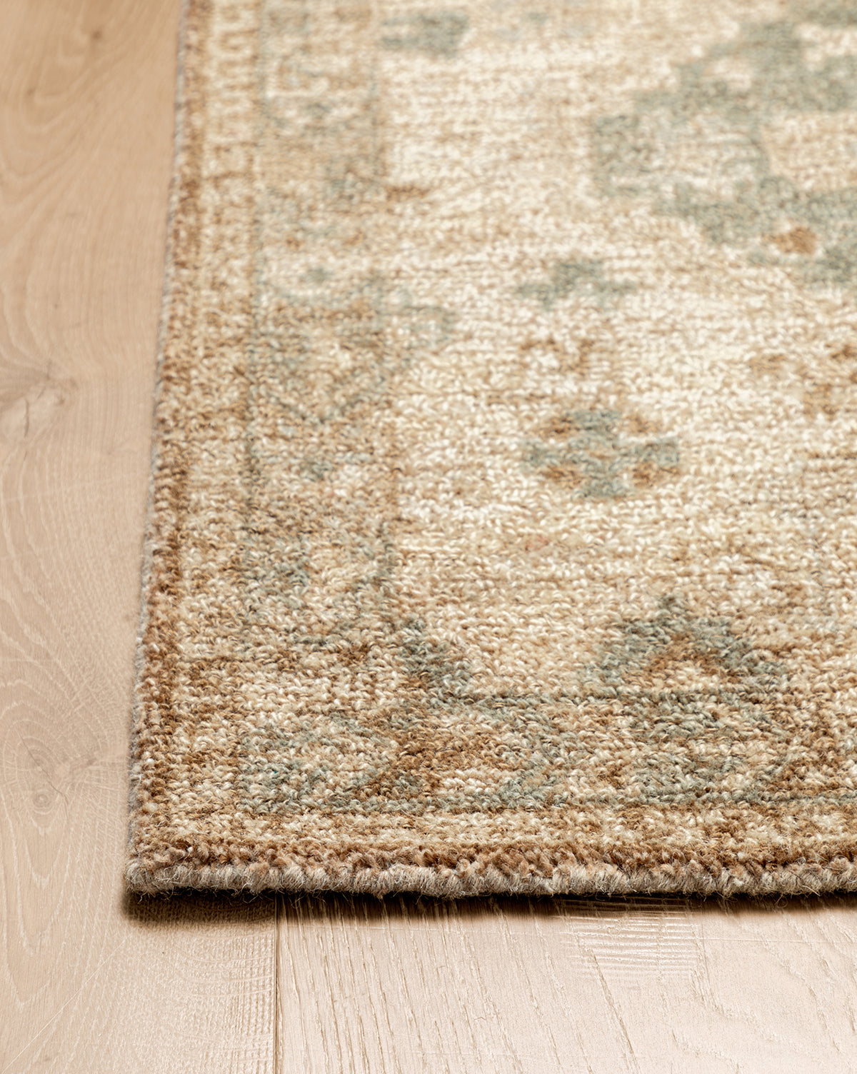 A close-up of the Wilshire Natural Hand-Tufted Wool Rug on a wooden floor highlights its intricate border, beige and light green pattern, textured wool surface, and spill-resistant qualities, by Vendixs