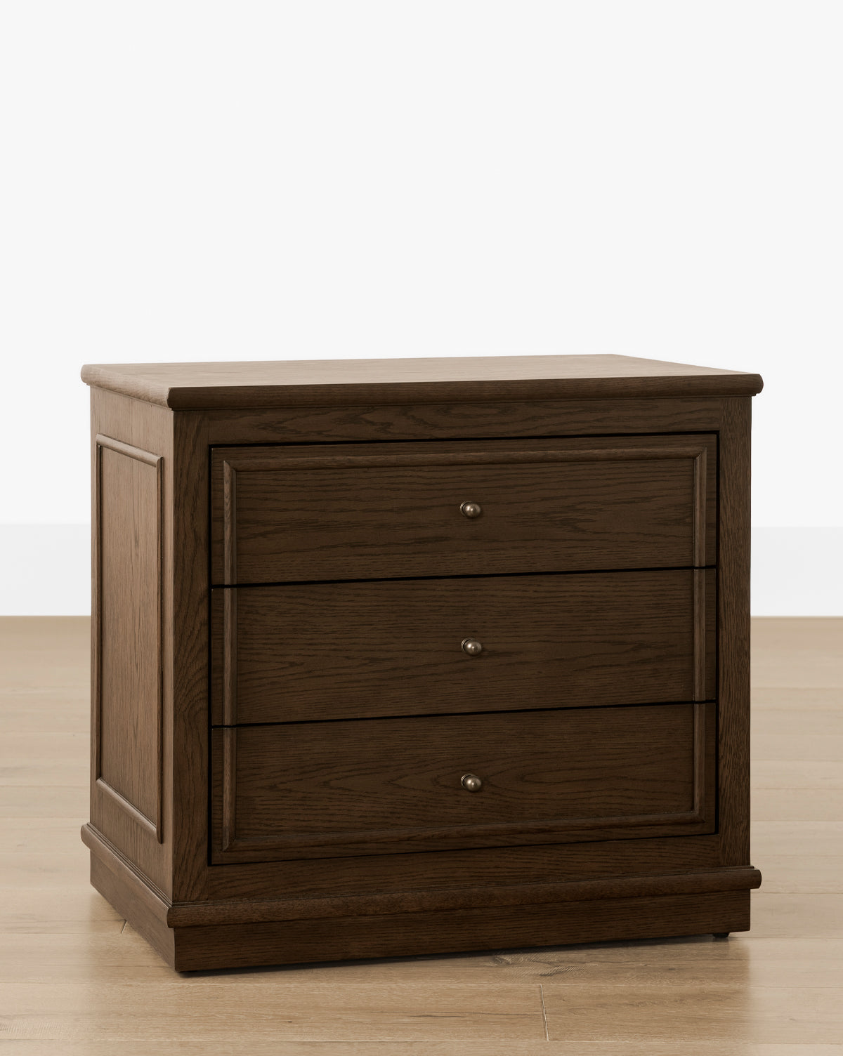 The Yorkshire Nightstand, finished in English oak, features three drawers with antique brass pulls and is displayed on a light wooden floor against a white background, by Vendixs