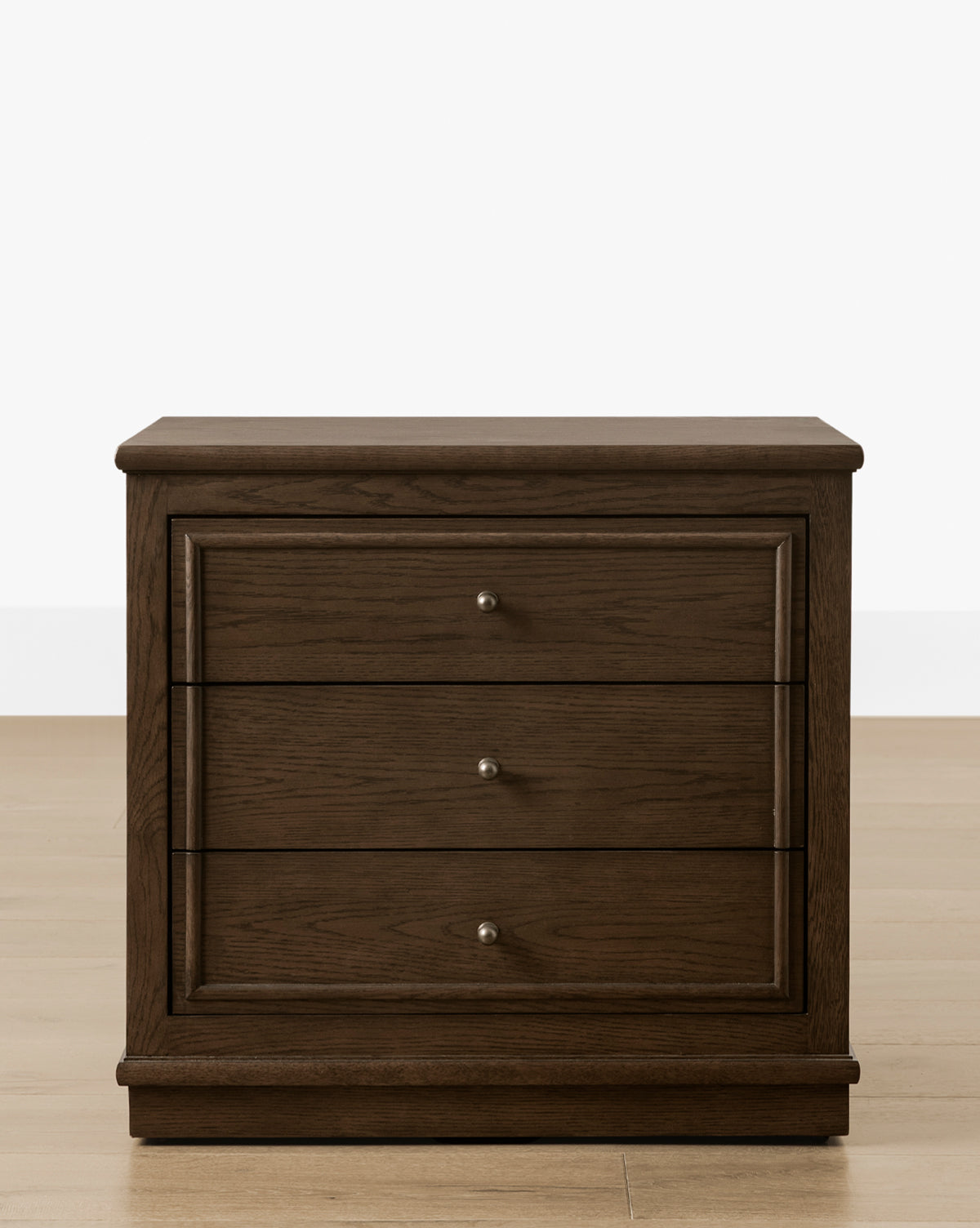 The Yorkshire Nightstand showcases a rich English oak finish with three drawers featuring antique brass pulls, elegantly placed on a light wood floor against a plain white background, by Vendixs