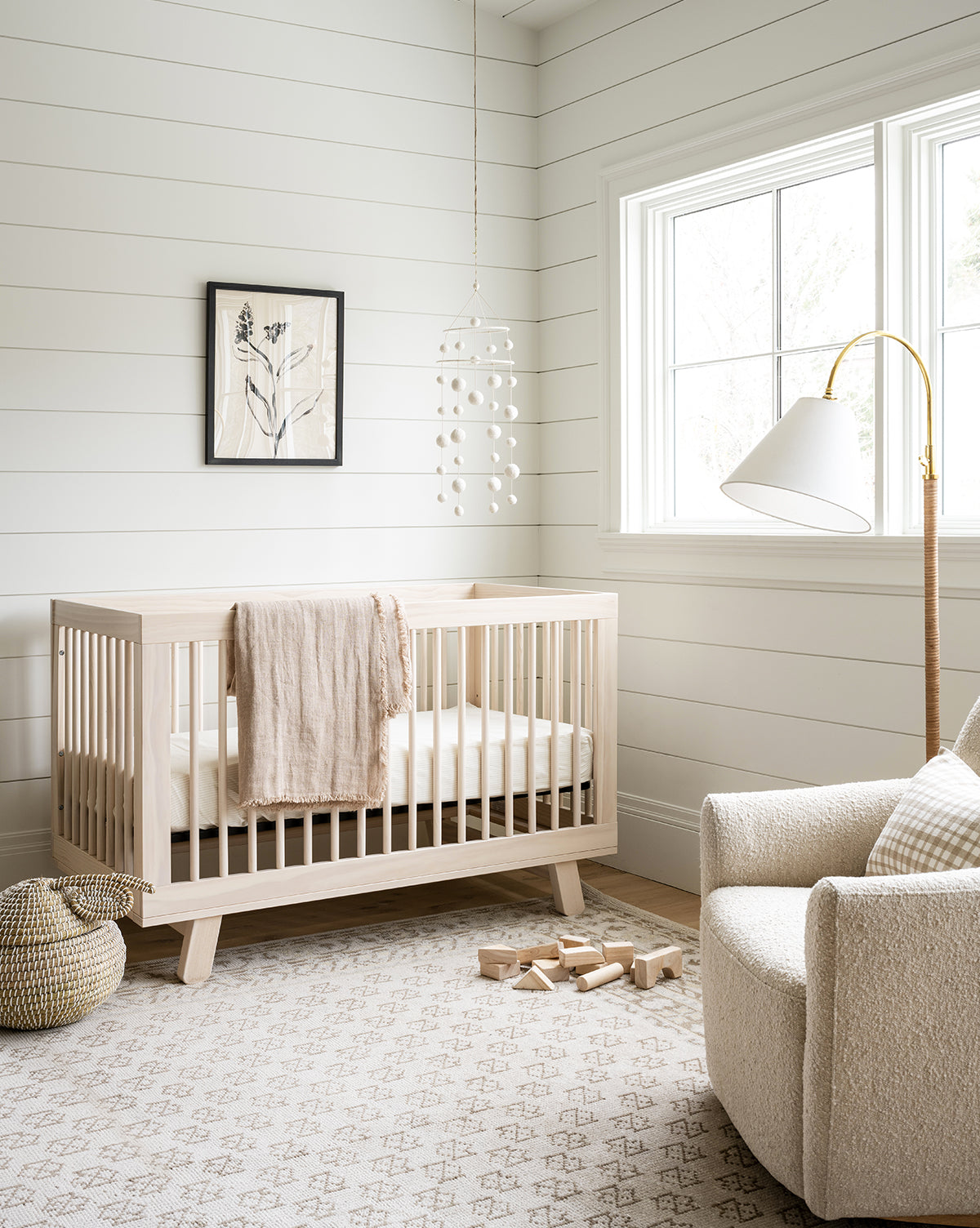 Bright, cozy nursery with a light wood crib, cream blanket, soft armchair, and the Anya Neutral Hand-Knotted Wool Rug. Wooden blocks, a basket, mobile, and botanical wall art add modern touches as sunlight streams through a large window, by Vendixs