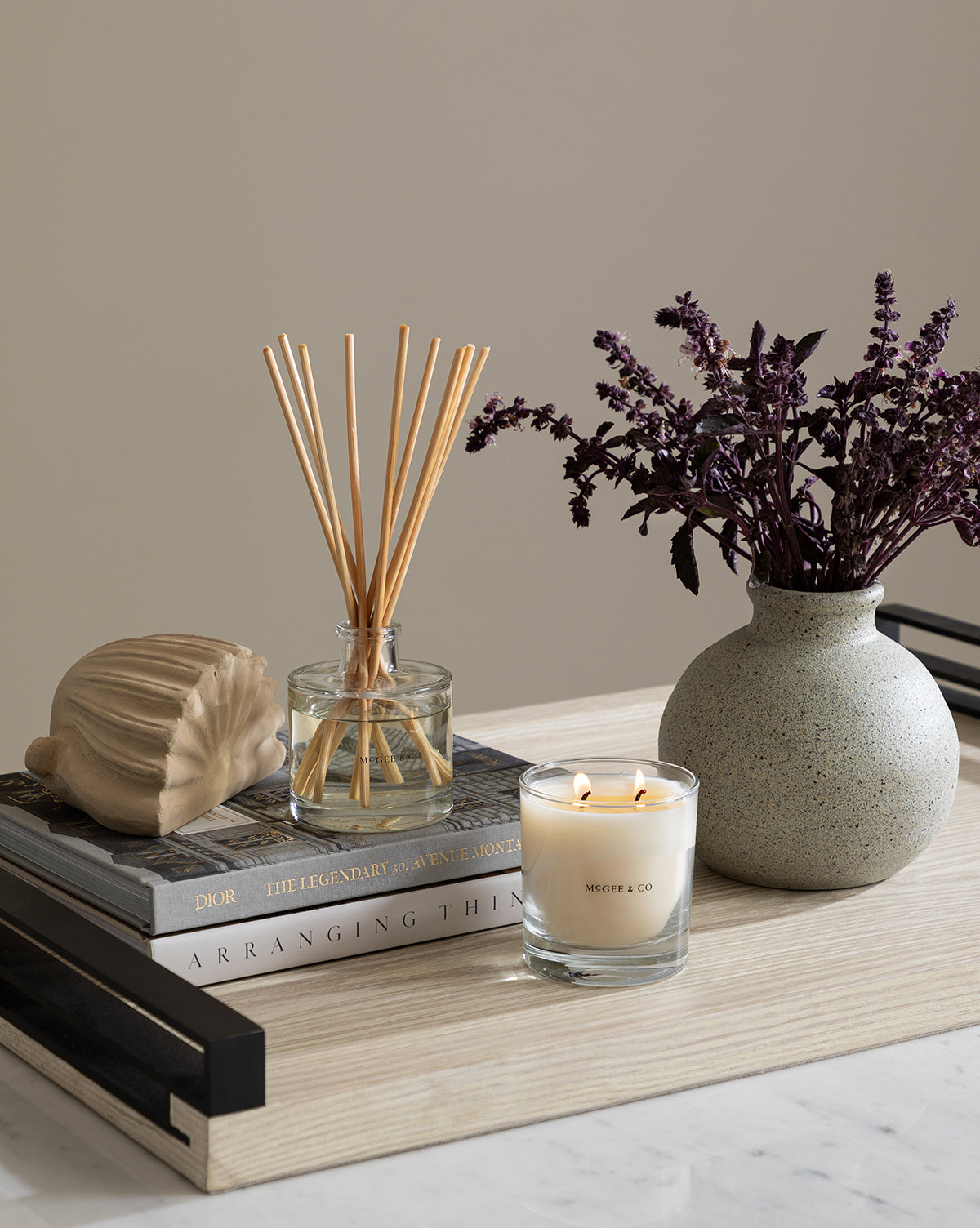 A Baroque Shell Object, candle, reed diffuser, and a textured vase with dark purple flowers are arranged on top of stacked books atop a light wooden surface.
