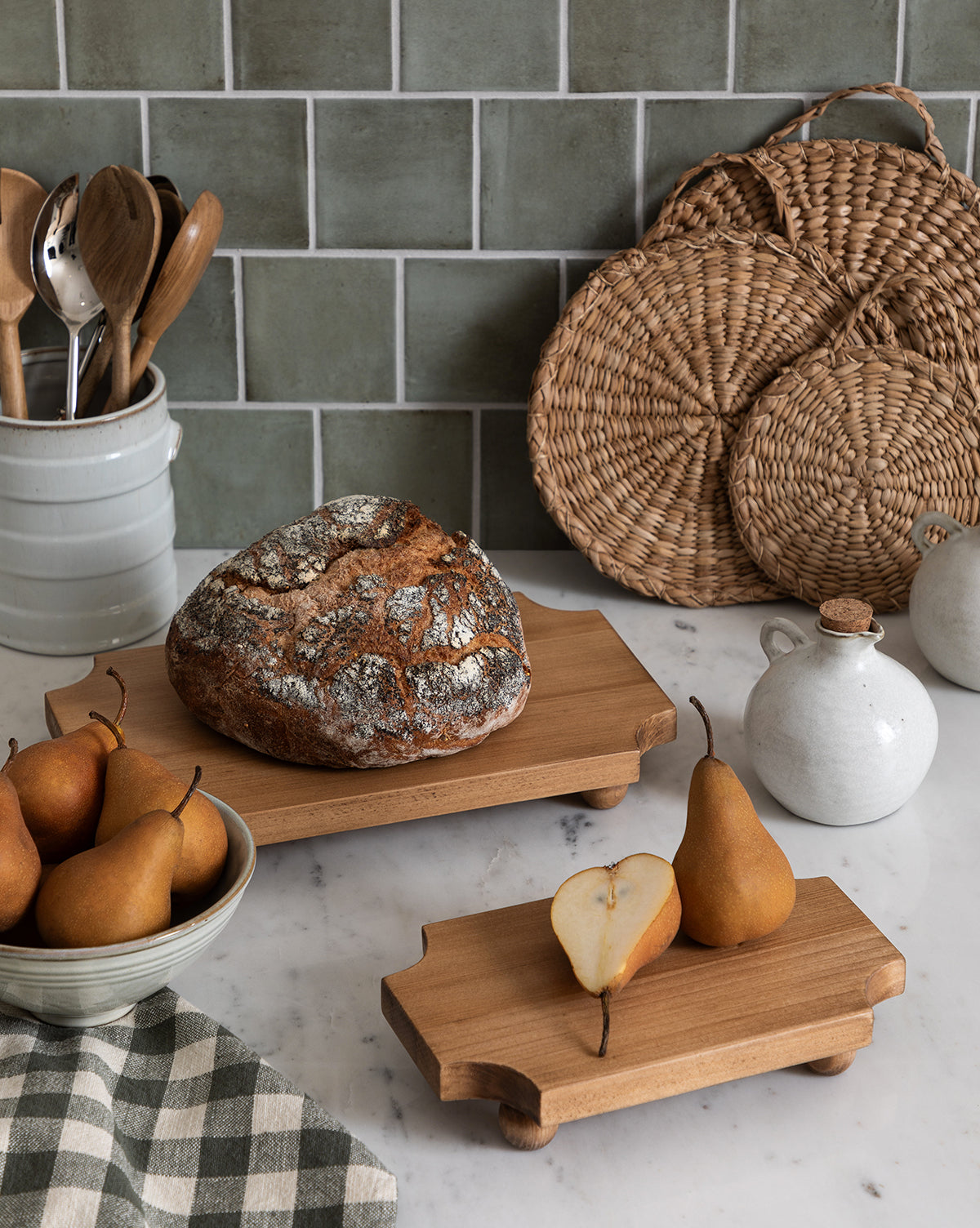 A rustic kitchen scene with pears in a bowl, a sliced pear on the Cillian Pedestal Board, a round loaf of bread, ceramic jars, wooden utensils in a crock, woven placemats, and a checkered cloth on a marble countertop by Vendixs