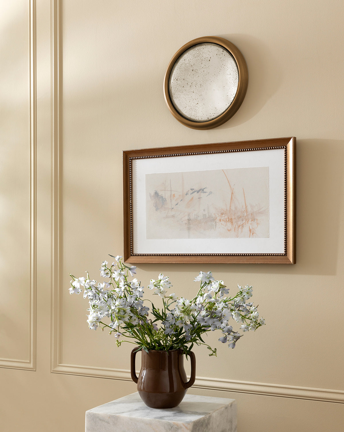 The Victoire Vase, in brown ceramic with white flowers, rests on a marble pedestal beneath a framed abstract artwork and a round decorative plate against a beige wall, by Vendixs
