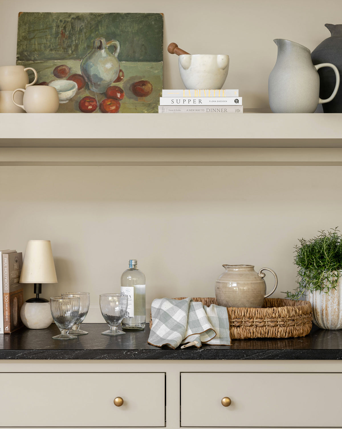 A kitchen countertop with glassware, a ceramic jug, folded checkered napkins, a potted plant, books, and a lamp. Above, a shelf displays pottery—including the Brown Stoneware Pitcher—books, and a still life painting with fruit and a white pitcher by Vendixs