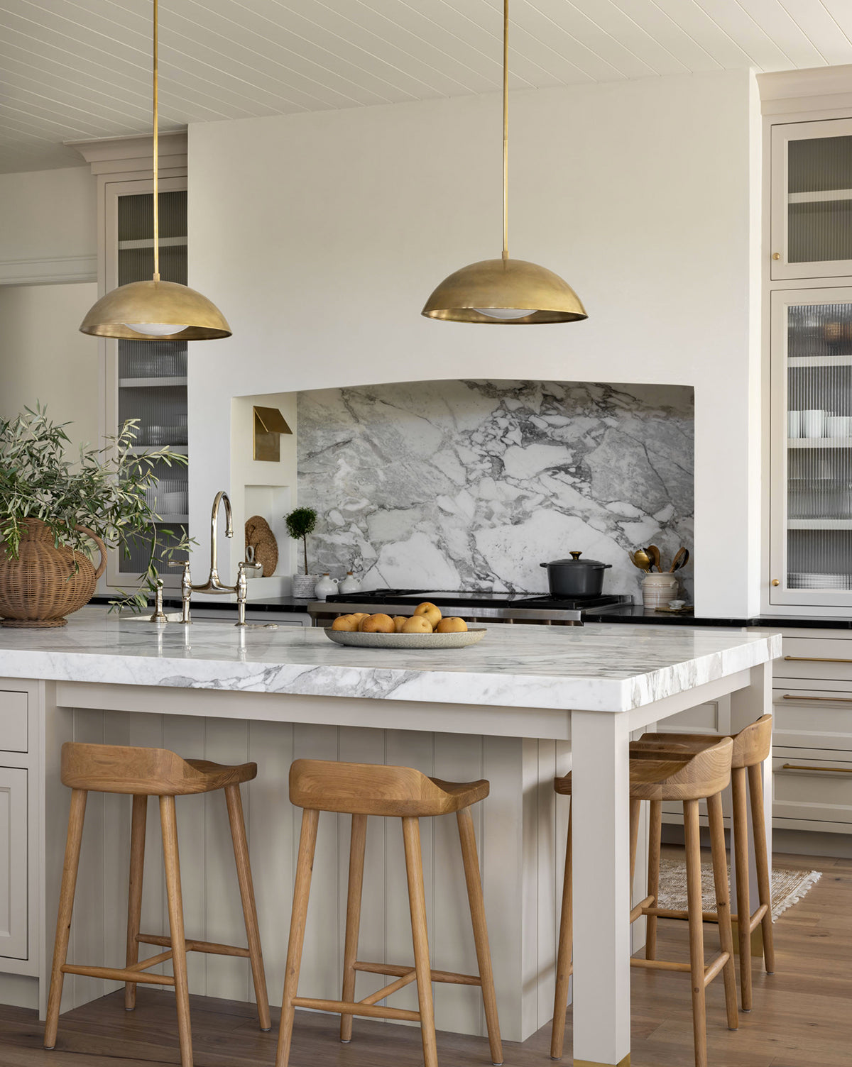 Modern kitchen with a marble island, Makerspalm Portland Counter Stools, gold pendant lights, and a marble backsplash. A bowl of lemons sits on the counter, while a potted plant accents the islands corner. Vendixs