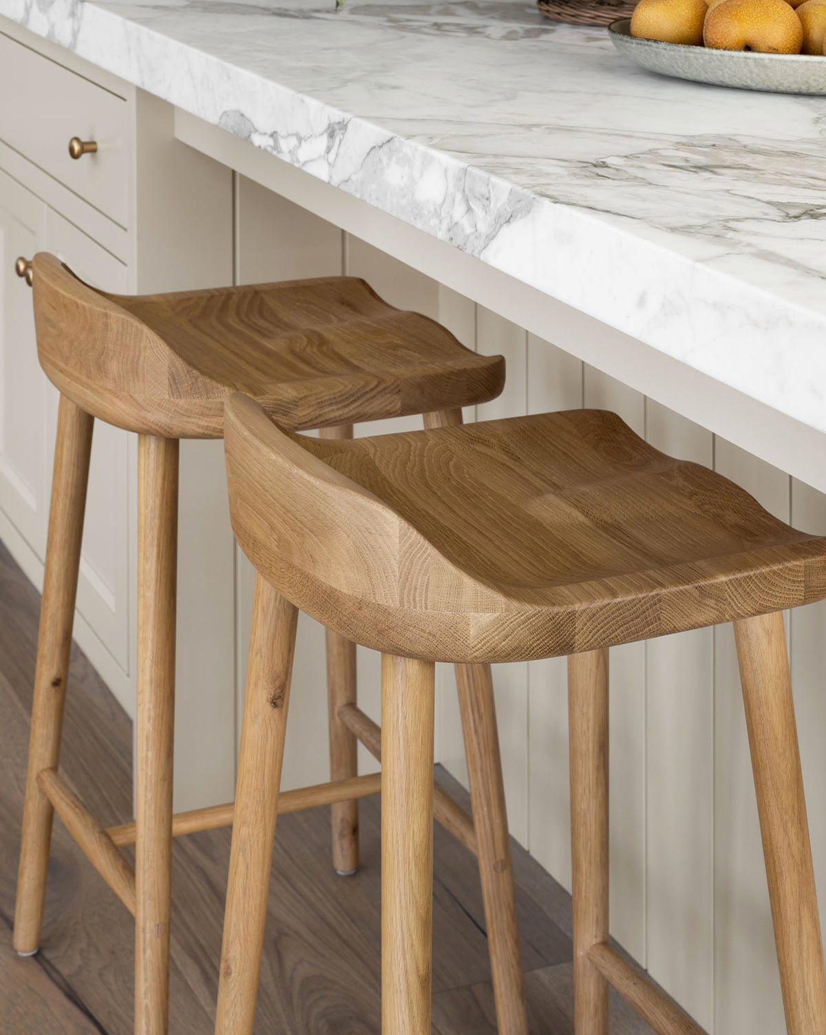 Two Makerspalm Portland Counter Stools with curved oak seats are set at a marble countertop in a modern kitchen with light cabinets and wood floors, Vendixs