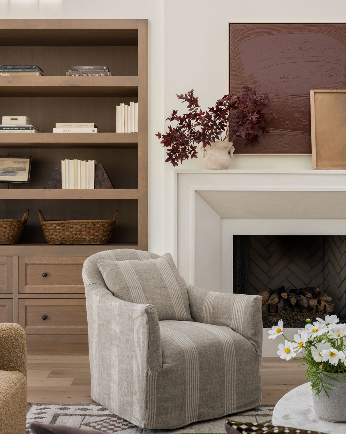The Trudeaux Slipcover Swivel Chair with gray and white stripes sits in front of a fireplace. Nearby, a wooden bookcase holds baskets and books, while a vase with red leaves and brown artwork decorate the mantel, by McGee & Co.
