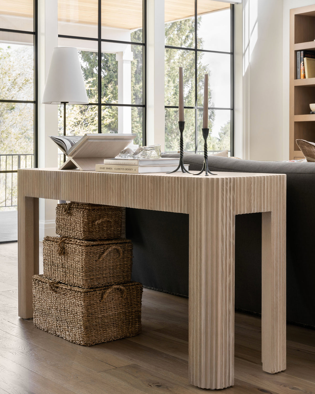 A light wood console table behind a dark sofa displays stacked books, Kya Seagrass Boxes, two tall candlesticks, and a white lamp. Three hand-woven baskets sit underneath. Large windows and shelves form the background by Vendixs