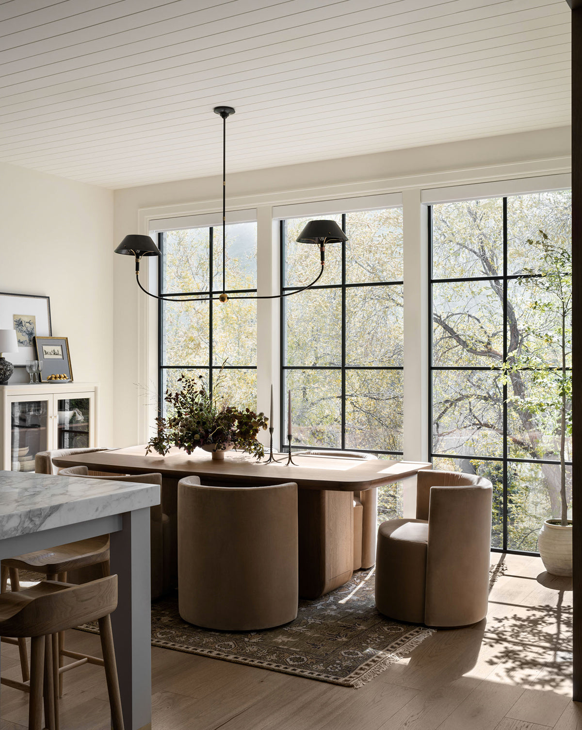 A sunlit dining area with large windows, a wooden table, round tan chairs, and a modern black chandelier highlights the Wayland Hand-Knotted Wool Rug, by Vendixs Decorative plants add charm beside a kitchen island with stools and a marble countertop.