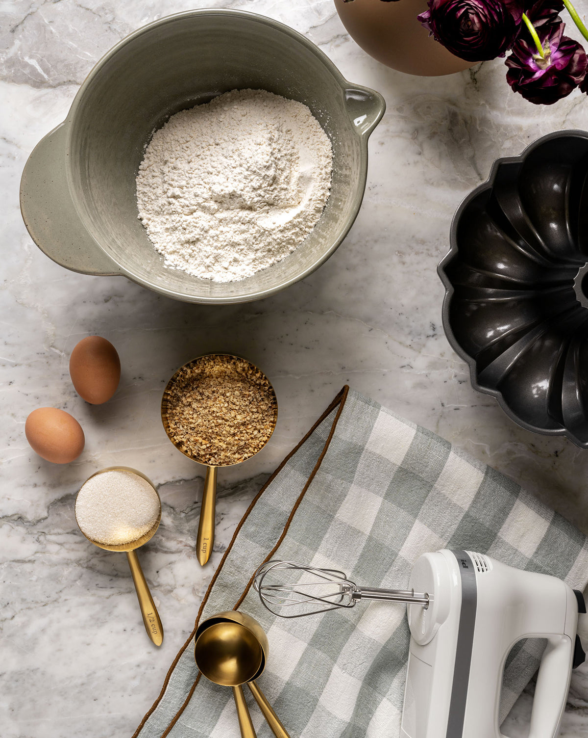 Baking essentials on a marble counter: a bowl of flour, two eggs, sugar and nuts in measuring cups, a Charlie Check Tea Towel, hand mixer, Bundt pan, and vase of dark flowers create a cozy table setting, by Vendixs