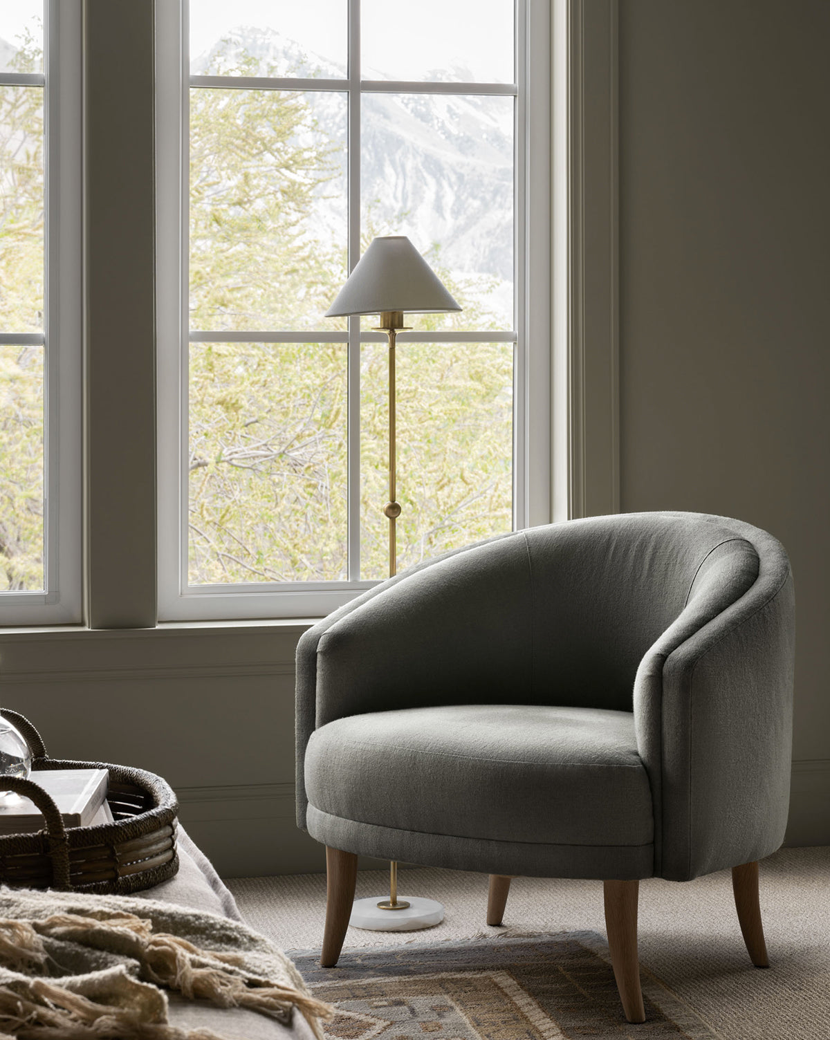The cozy Marguerite Lounge accent chair in gray sits by a large window with mountain and tree views, paired with a brass floor lamp and a woven basket nearby. Vendixs