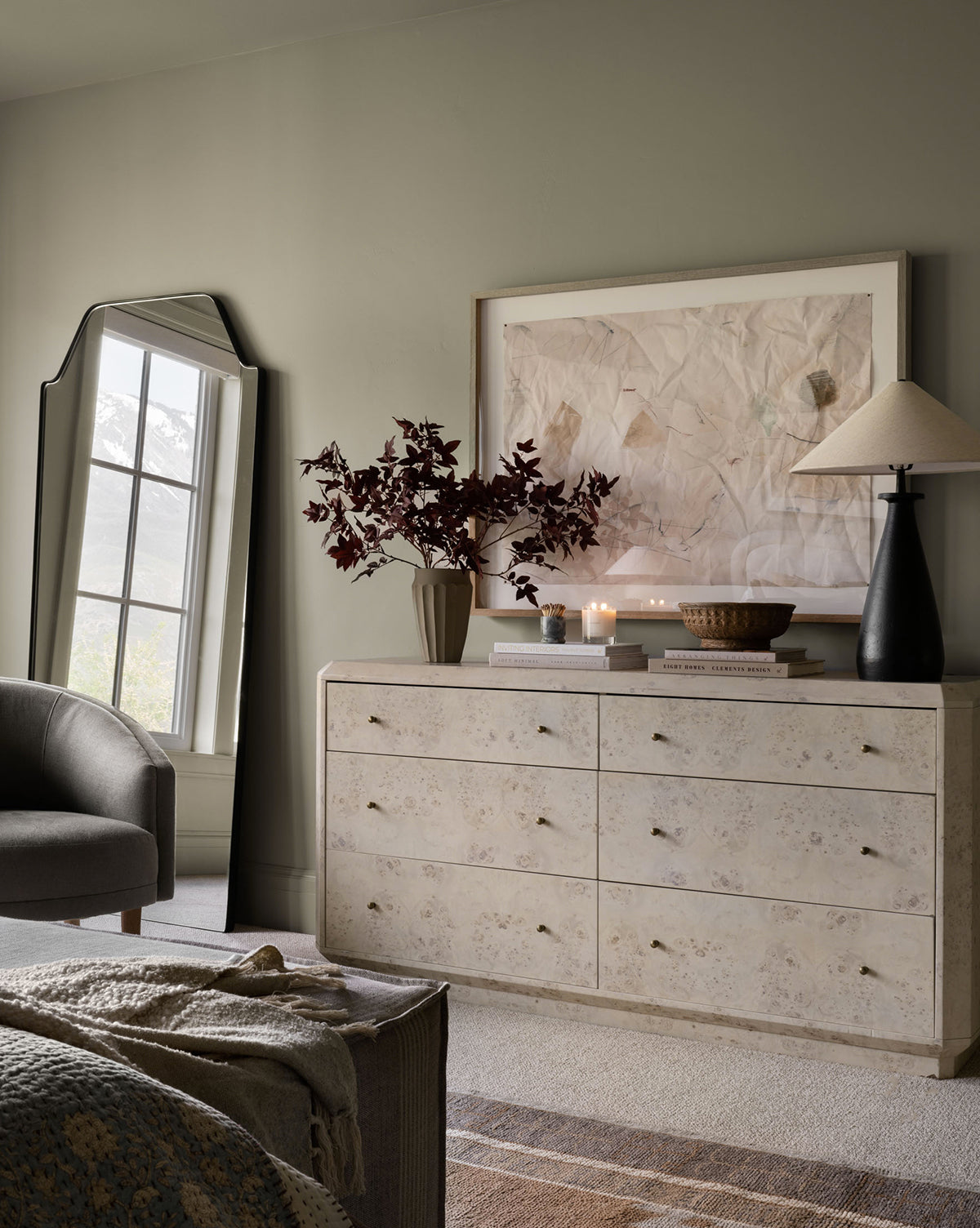 A light dresser with gold knobs displays a vase with dark red leaves, candles, a basket, and two lamps. The Giles Floor Mirror with a matte black frame leans against the wall by the window; abstract art is framed above the dresser, by Vendixs