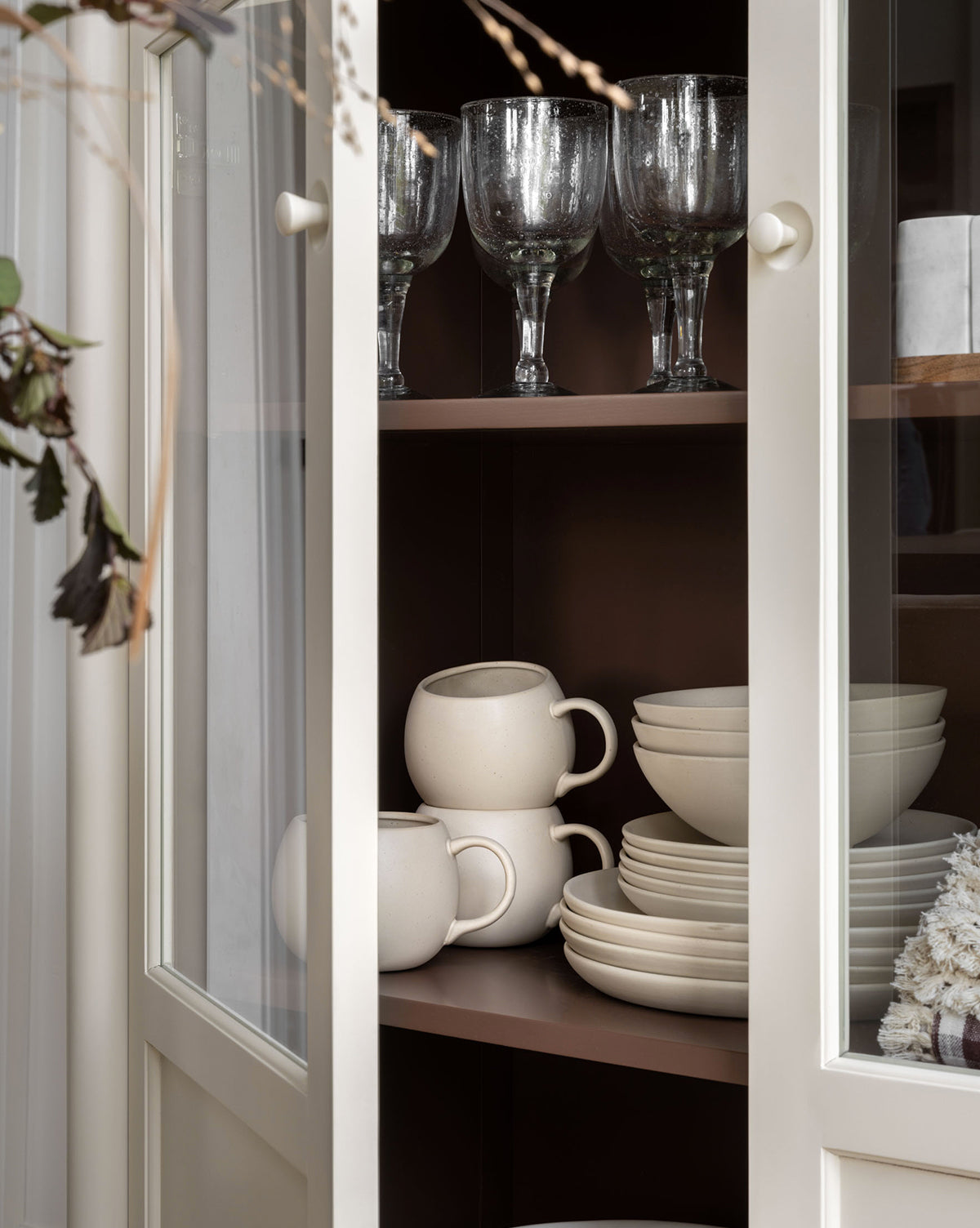 A cream-colored cabinet with glass doors displays stacked white ceramic cups and plates, including the Luana Bowl on the bottom shelf, and clear goblets above. The modern neutral scene features dried foliage to the left, by Vendixs