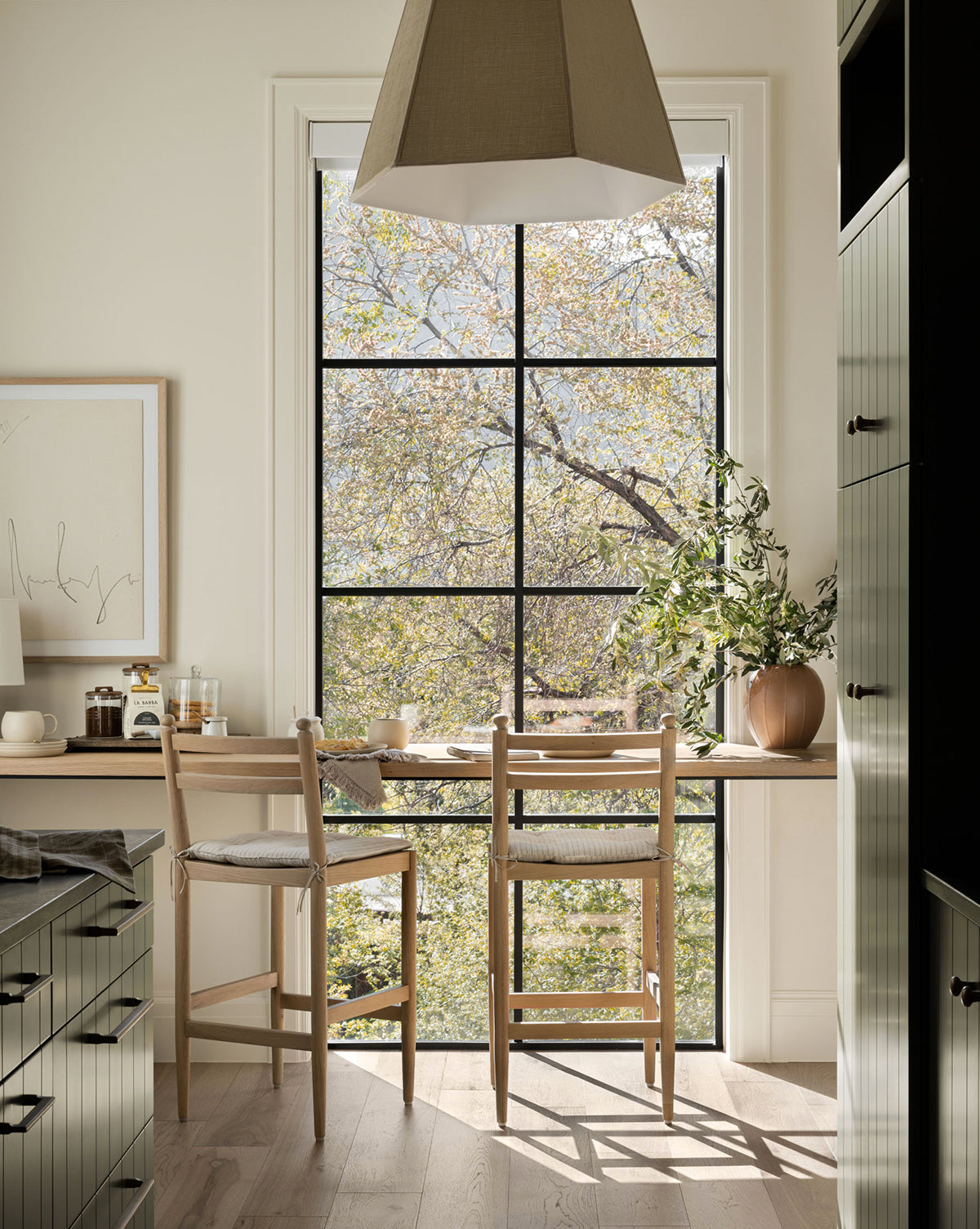 A bright kitchen nook features two Romany Counter Stools with linen cushions by a large window overlooking leafy trees. Natural light fills the space, while plants and simple decor create a cozy, inviting atmosphere by Vendixs