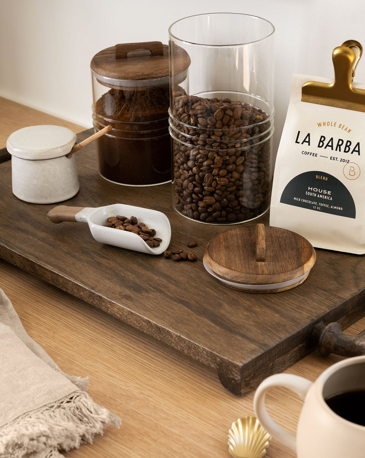 The Fordham Board displays coffee beans in glass jars, ground coffee in a jar, a “La Barba Coffee” bag, a ceramic scoop with beans, a cup of black coffee, and a linen napkin on a wooden surface, by Vendixs