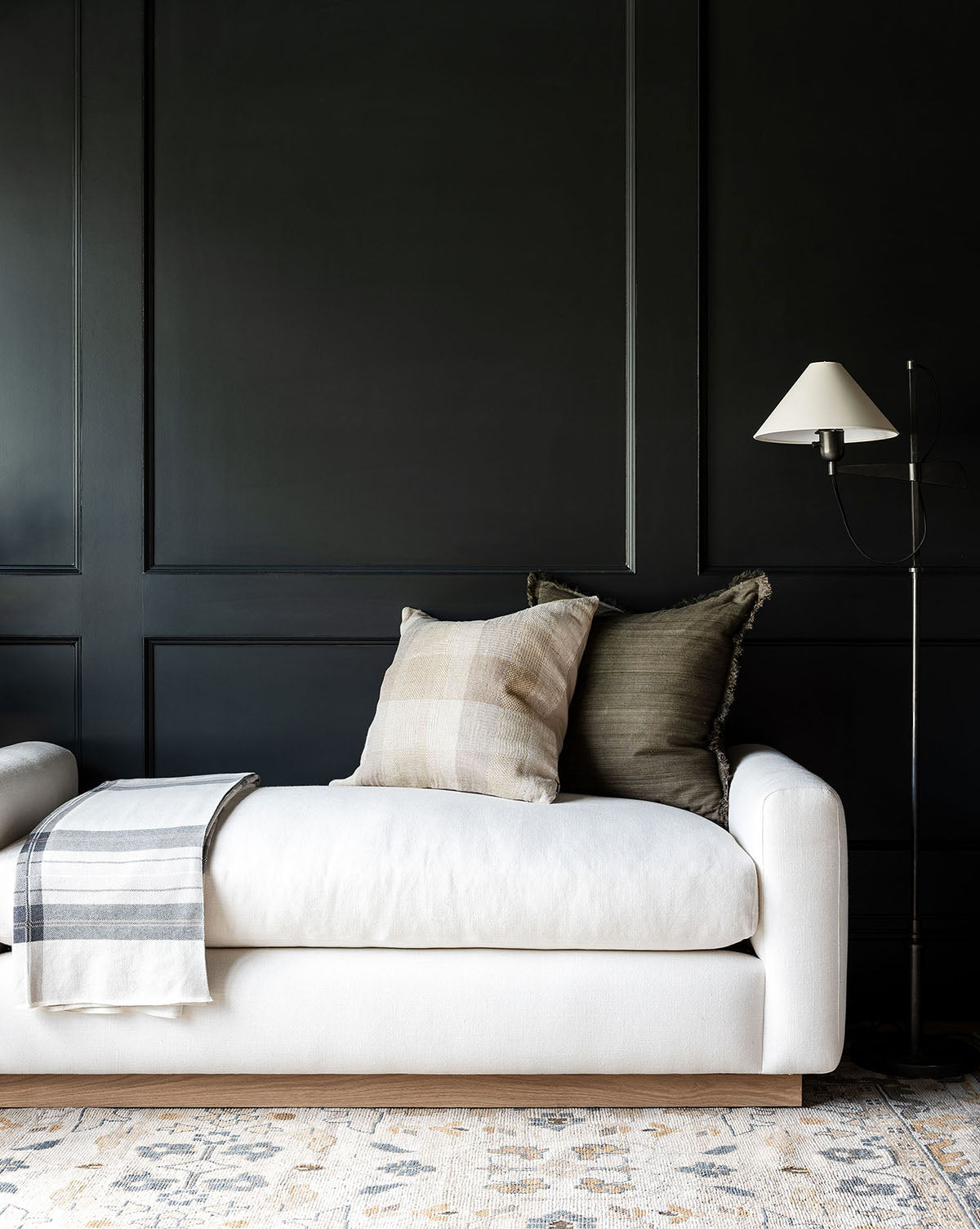 A modern living room features a white sofa, neutral pillows, a striped blanket, patterned rug, and black paneled wall. The Mansfield Bench sits nearby, providing stylish extra seating and enhancing the space’s contemporary look by McGee & Co.