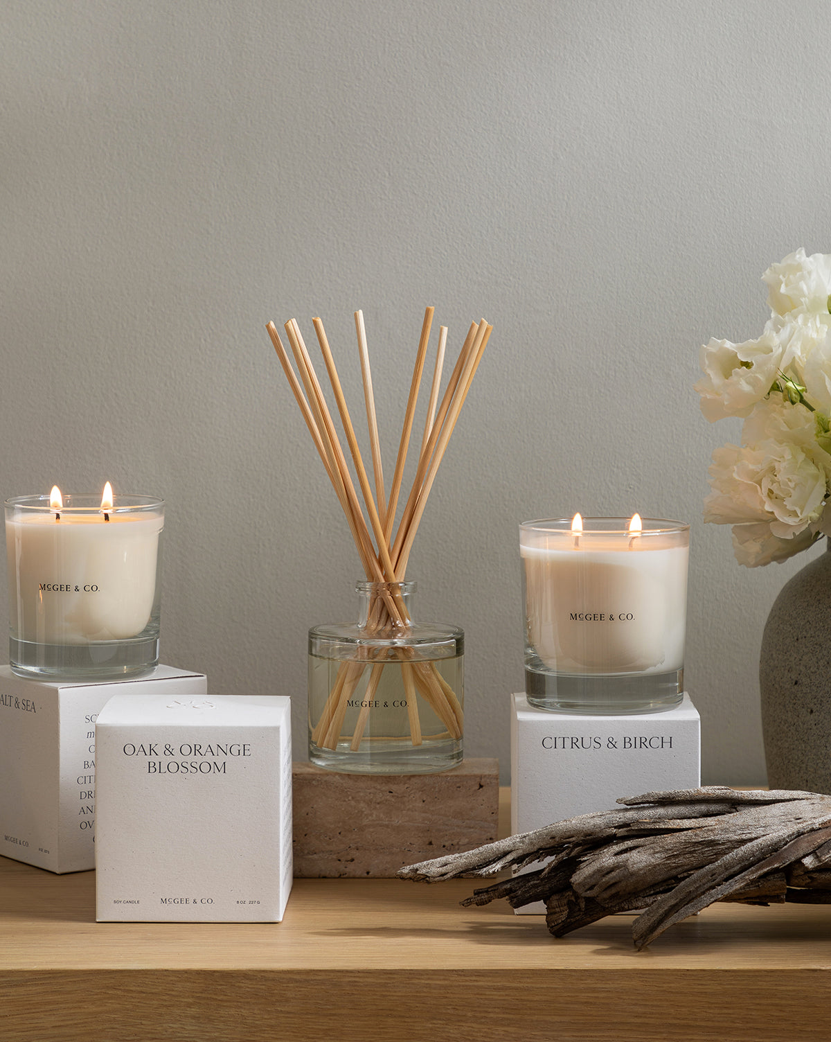 A minimalist display with two lit soy candles, a glass diffuser, white flowers in a vase, driftwood, and luxury candle boxes—including one labeled Citrus & Birch Candle—all arranged on a light wooden surface, by Vendixs