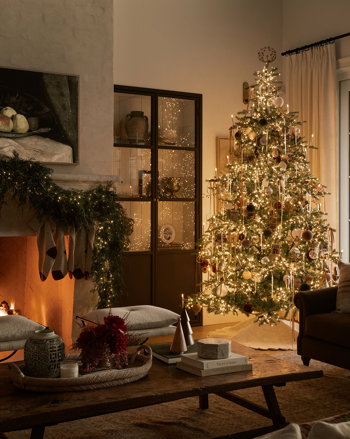 A warmly lit living room decorated for Christmas features a glowing 9' Grandis Fir Pre-Lit Faux Tree with ornaments, stockings on a garlanded fireplace, and cozy cushions with books atop a wooden coffee table, by Vendixs