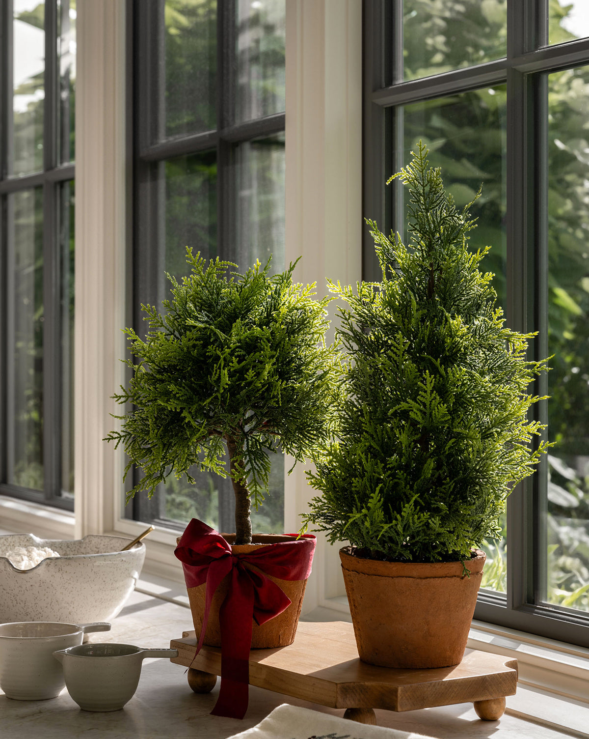 Two 15.5" Cypress Faux Potted Topiary trees sit on a sunlit windowsill. One terracotta pot has a red ribbon, and both are on a wooden tray. Kitchen bowls are nearby, with greenery visible outside through the window, by Vendixs