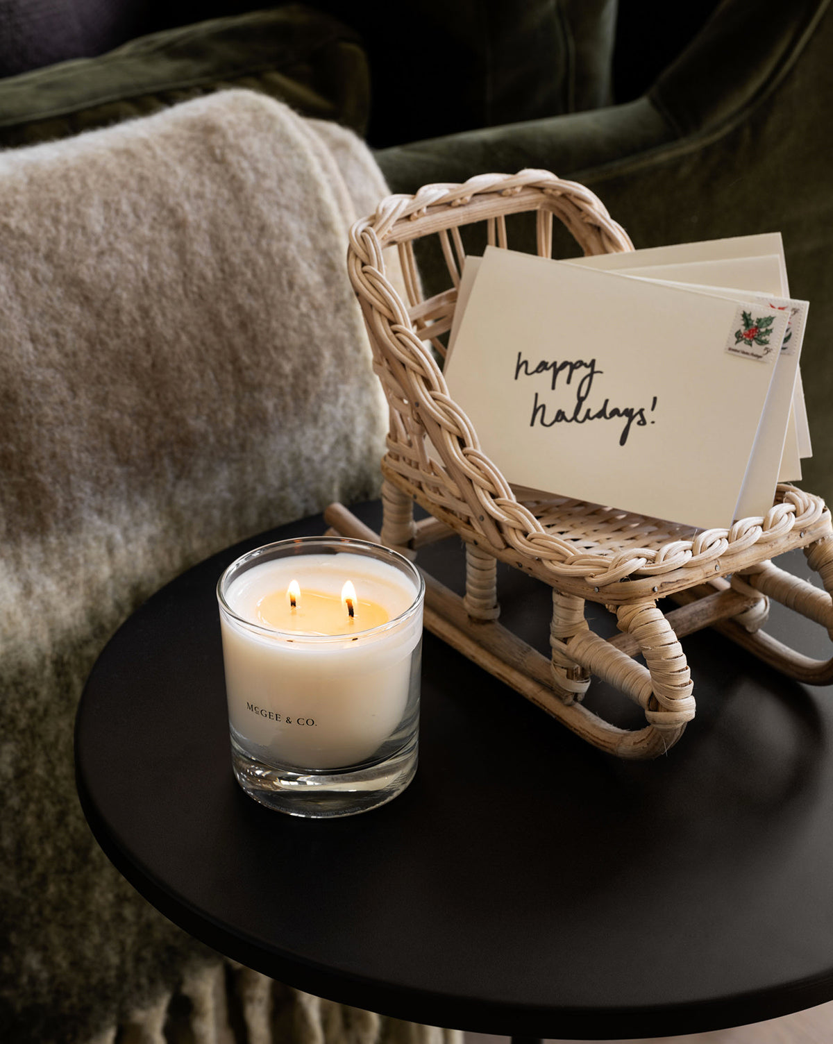 A glass candle with two flames sits on a black table beside the Woven Rattan Sleigh in a natural finish, holding holiday cards—one says “happy holidays!” A textured blanket and green cushion complete the festive decor by Vendixs