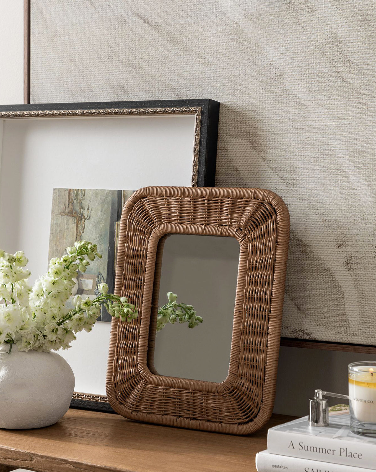 The Jera Shelf Mirror, a rectangular woven rattan piece, rests on a wooden surface beside a white flower vase, framed art print, candle, and book. Behind them hangs a large light-toned textured artwork, by Vendixs