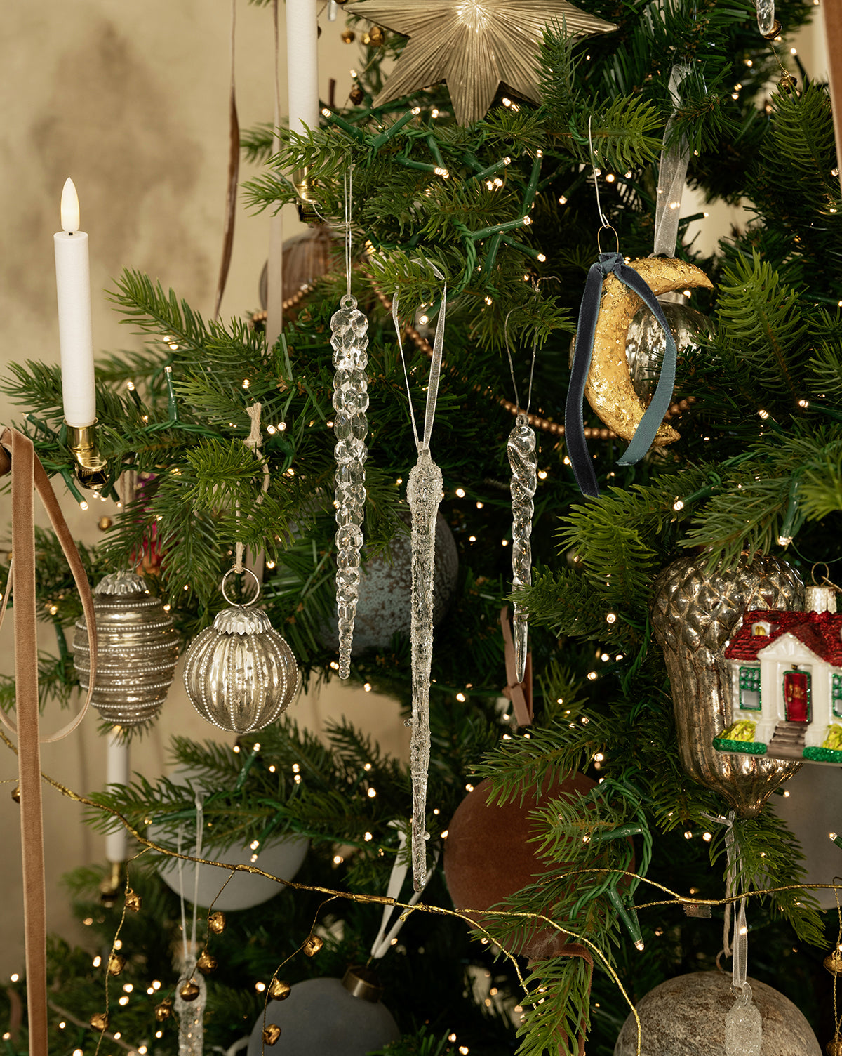 A Christmas tree adorned with glass ornaments, a set of 3 Frosted Icicle Ornaments, silver baubles, a candle-shaped ornament, a star topper, bird and house ornaments, all glowing under twinkling string lights, by Vendixs