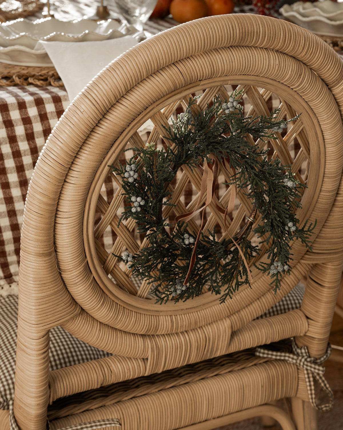 A woven rattan chair featuring the 8" Blueberry & Juniper Faux Wreath hangs beside a table draped in a brown and white checkered cloth, set with plates, napkins, and glasses—ideal for cozy holiday decor, by Vendixs