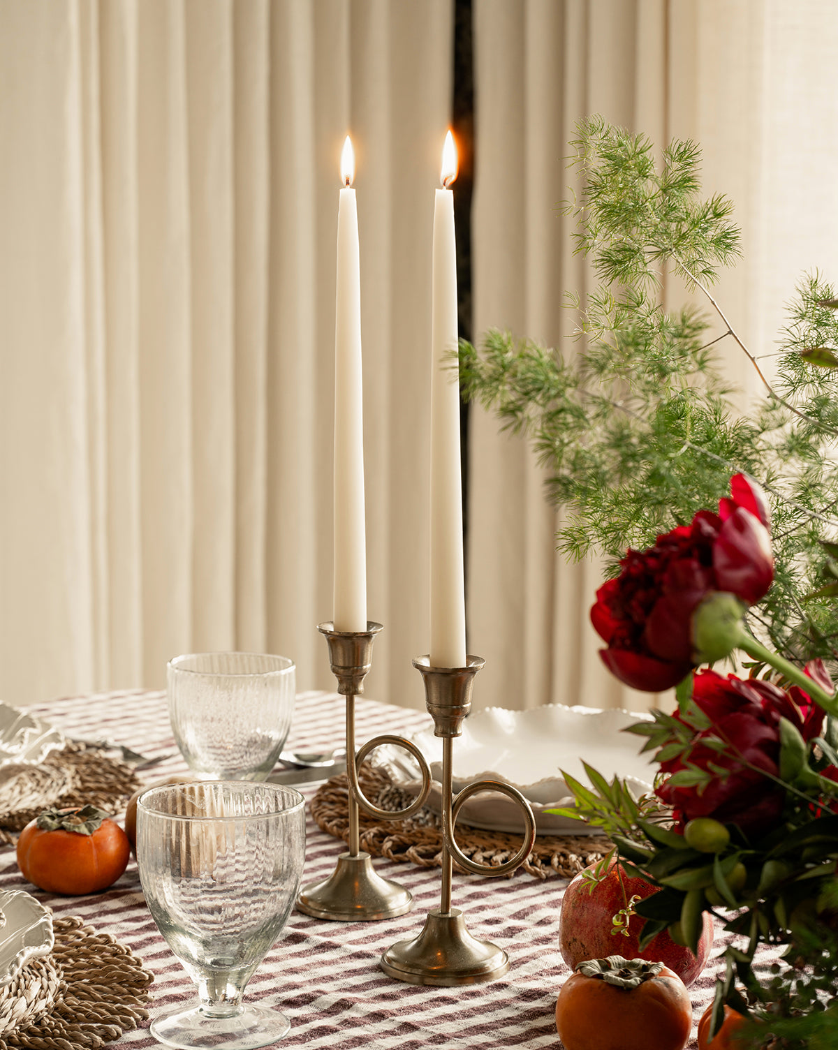 A dining table set with two lit candles in French Horn Taper Holders, clear glasses, woven placemats, red flowers, greenery, persimmons, and a pomegranate on a checkered tablecloth—ideal for elegant holiday decor, by Vendixs