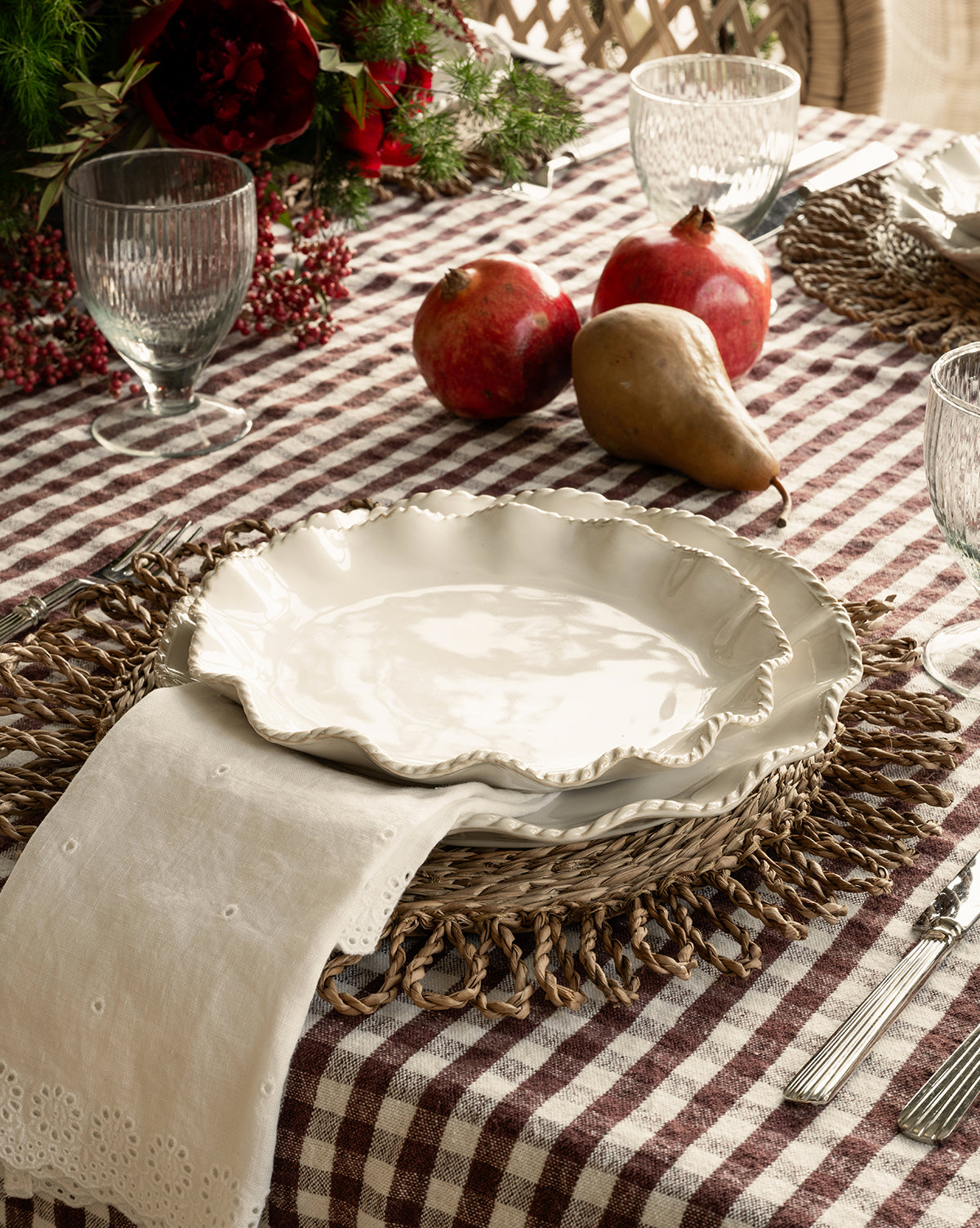 A rustic table with white ceramic plates on woven chargers, a checkered cloth, glassware, silverware, two pomegranates, a pear, greenery with red berries, and Vendixs x Loeffler Randall Delilah Broderie Napkins by McGee & Co.