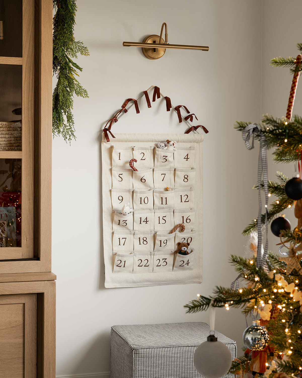 An Advent Calendar with numbered pockets hangs on the wall, decorated with small gifts. Nearby, a lit Christmas tree and festive greenery on a cabinet create a cozy holiday atmosphere, by Vendixs