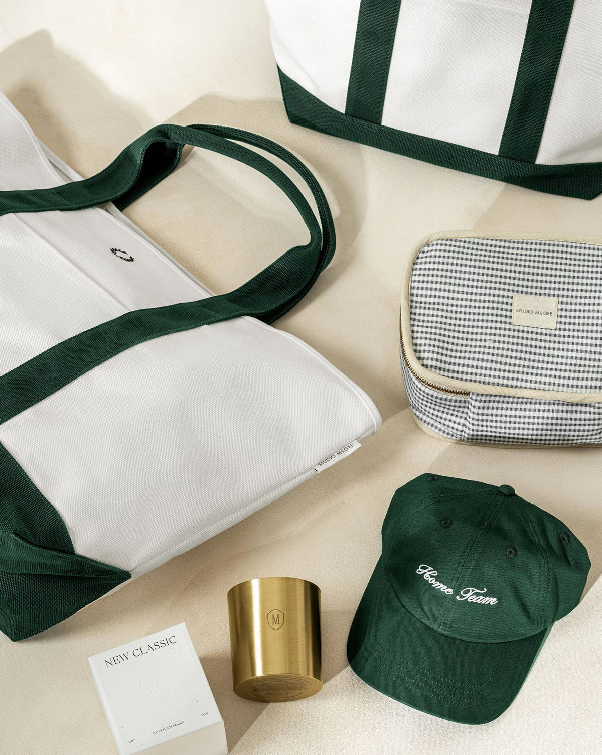 A flat lay of stylish items on a light surface, featuring two white and green tote bags, a green cap, a small checkered pouch, an Embossed Brass Candle in a gold vessel, and a white boxed perfume, by Vendixs