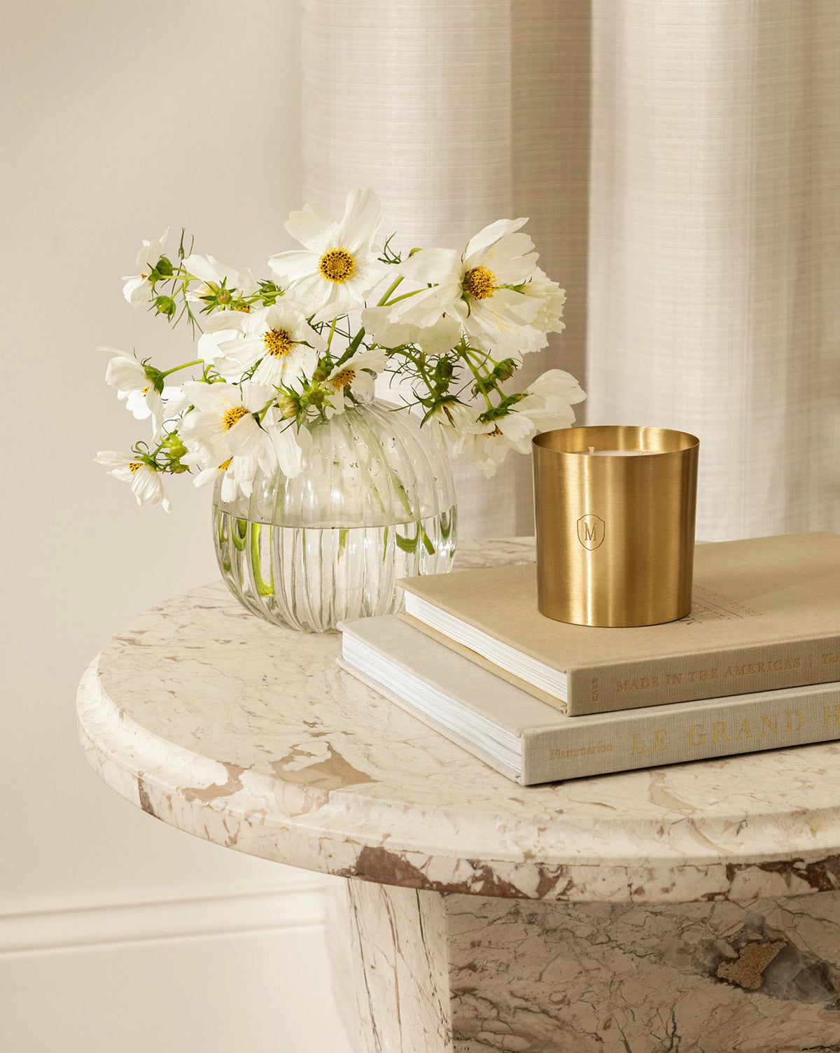 A round marble table features an Embossed Brass Candle, a glass vase of white flowers, a gold cup, and two stacked books; soft light-colored curtains hang in the background, by Vendixs