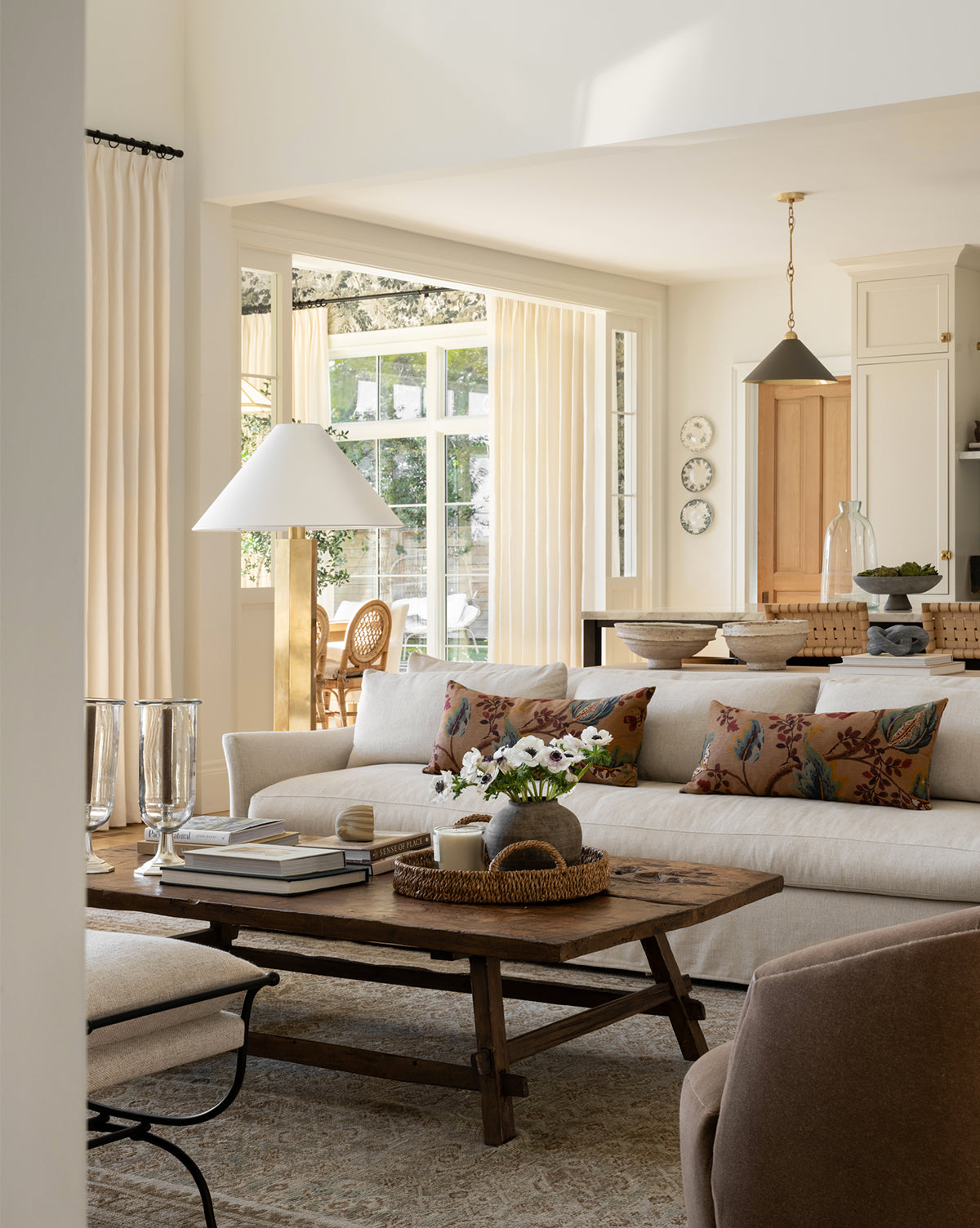 A cozy, sunlit living room features the Wilhelmina Classic Slipcover Sofa (85-110) in beige with floral pillows, a wooden coffee table topped with books and flowers, cream-curtained windows, and a dining area in the background by Vendixs