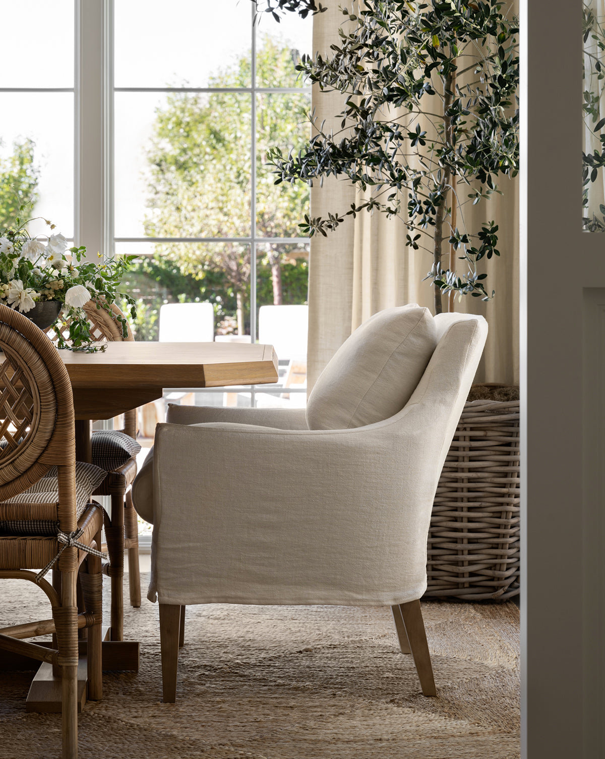 A cozy dining room featuring the Sascha Slipcover Dining Chair by Rowe Fine Furniture, Inc., a wood dining table, woven chairs, a large wicker basket, leafy indoor plant, and sunlight pouring through windows with garden views—McGee & Co.