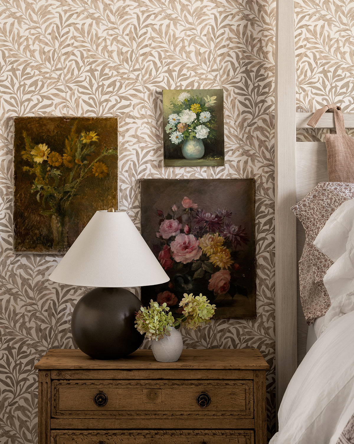 A cozy bedroom corner features the Tess Ceramic Table Lamp on a wooden nightstand, with a small flower vase and three floral paintings set against leafy wallpaper beside a white bed, by Vendixs