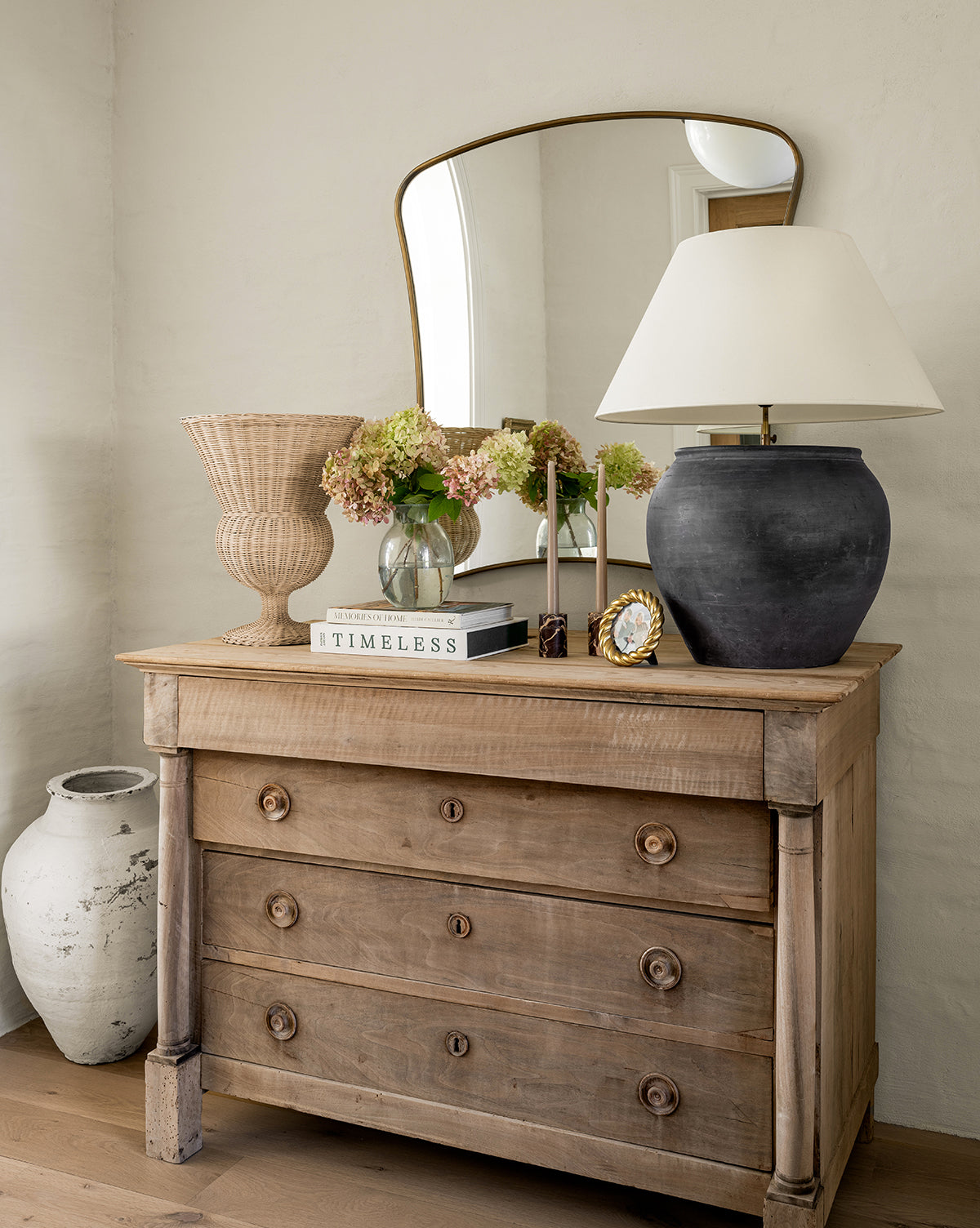 A rustic wooden dresser with three drawers displays a black lamp, wicker basket, floral vase, books, gold clock, and the Ludwig Wall Mirror. Beside the dresser on the floor is a large decorative ceramic pot, by Vendixs