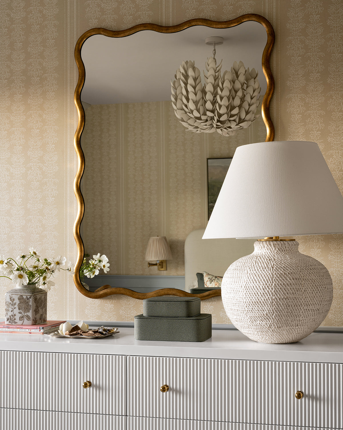 A dresser with gold knobs displays the Avedon Table Lamp with a white linen shade, two green boxes, and a vase of white flowers. Above, a large wavy gold-framed mirror reflects light fixtures and part of the room, by Vendixs