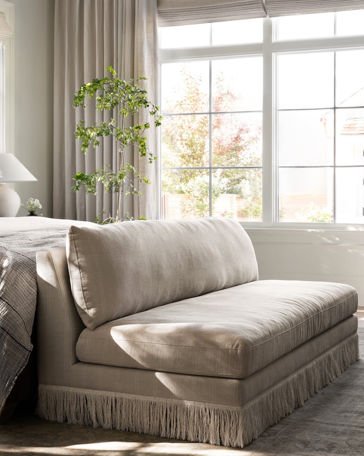 The Philippa Settee (Ready to Ship) with fringe trim sits by a sunlit window, next to a neatly made bed and tall potted plant, creating a cozy, serene bedroom corner, by Vendixs