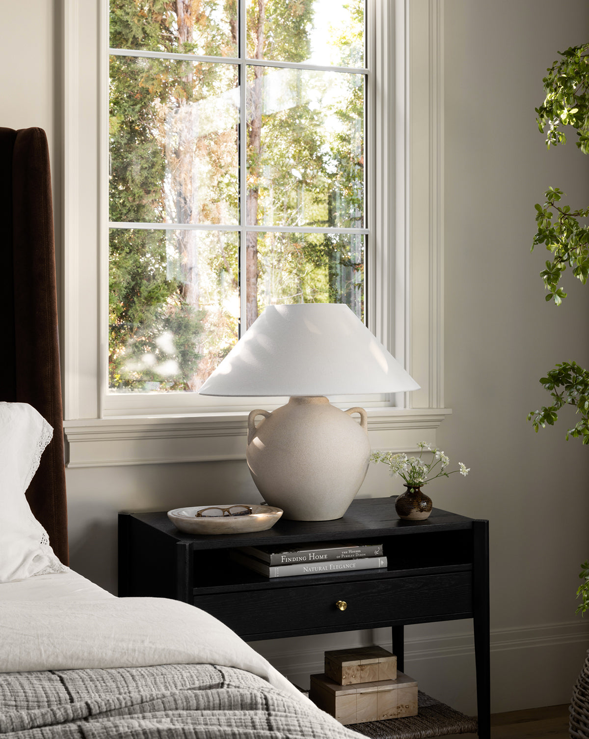 A cozy bedroom scene features a neatly made bed next to a black nightstand topped with the Mays Table Lamp, a dish, stacked books, and a small vase of flowers, all set before a sunlit window overlooking trees by Vendixs