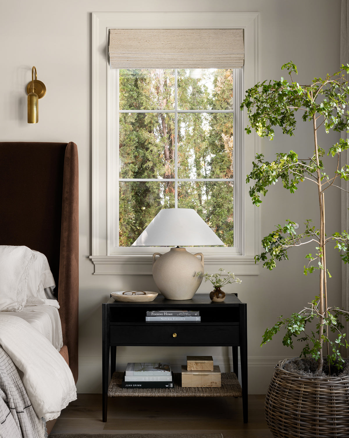 The Dana Nightstand, adorned with books, a ceramic vase, and a lamp, stands beside a bed with white bedding. Behind it is a window revealing green trees. To the right is a large potted plant; to the left hangs a brass wall sconce, by McGee & Co.