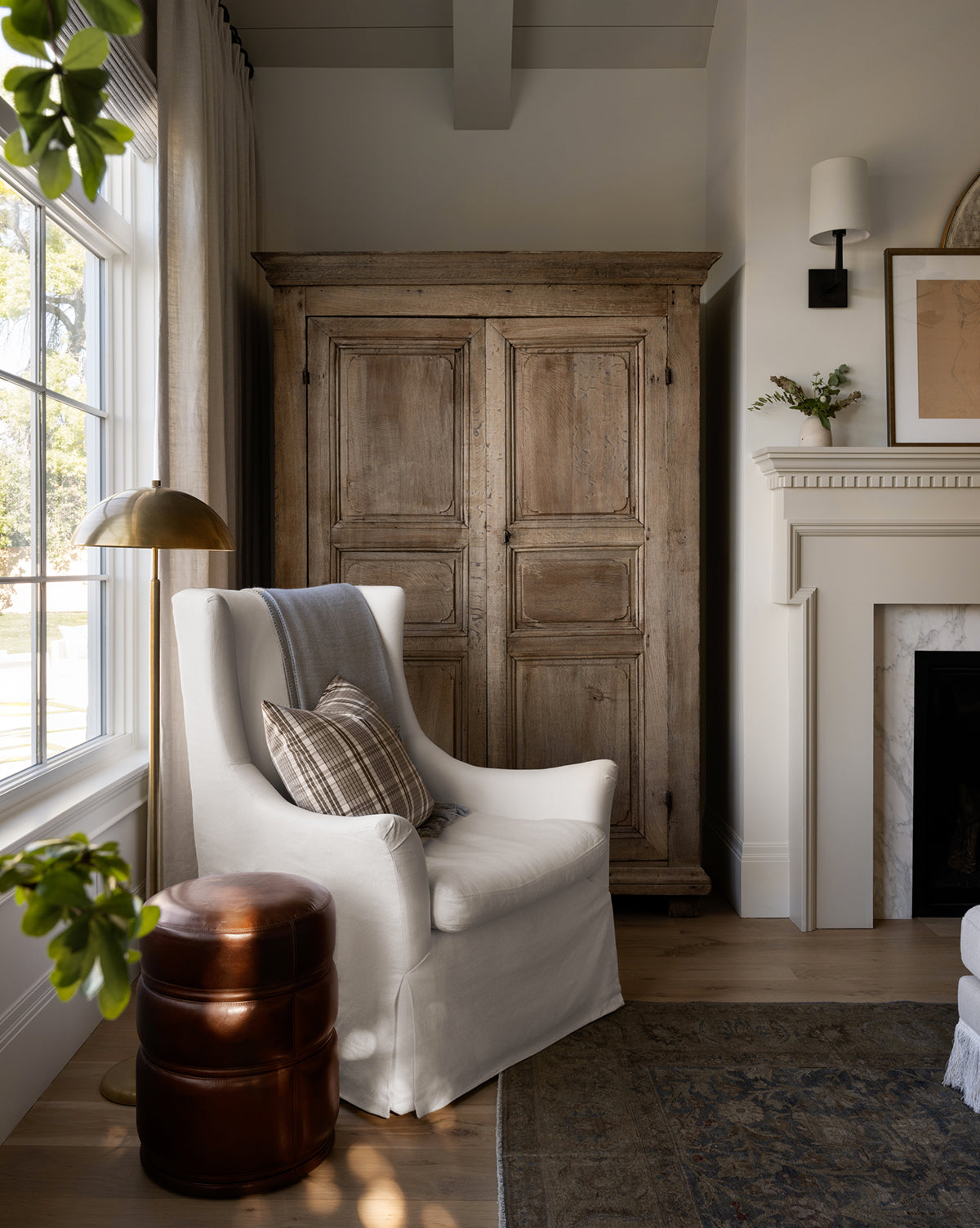 A cozy corner features a white armchair, plaid and striped pillows, a brown leather ottoman, the elegant Aldorno Floor Lamp, a large wooden armoire, window, and a white fireplace adorned with decor, by Vendixs