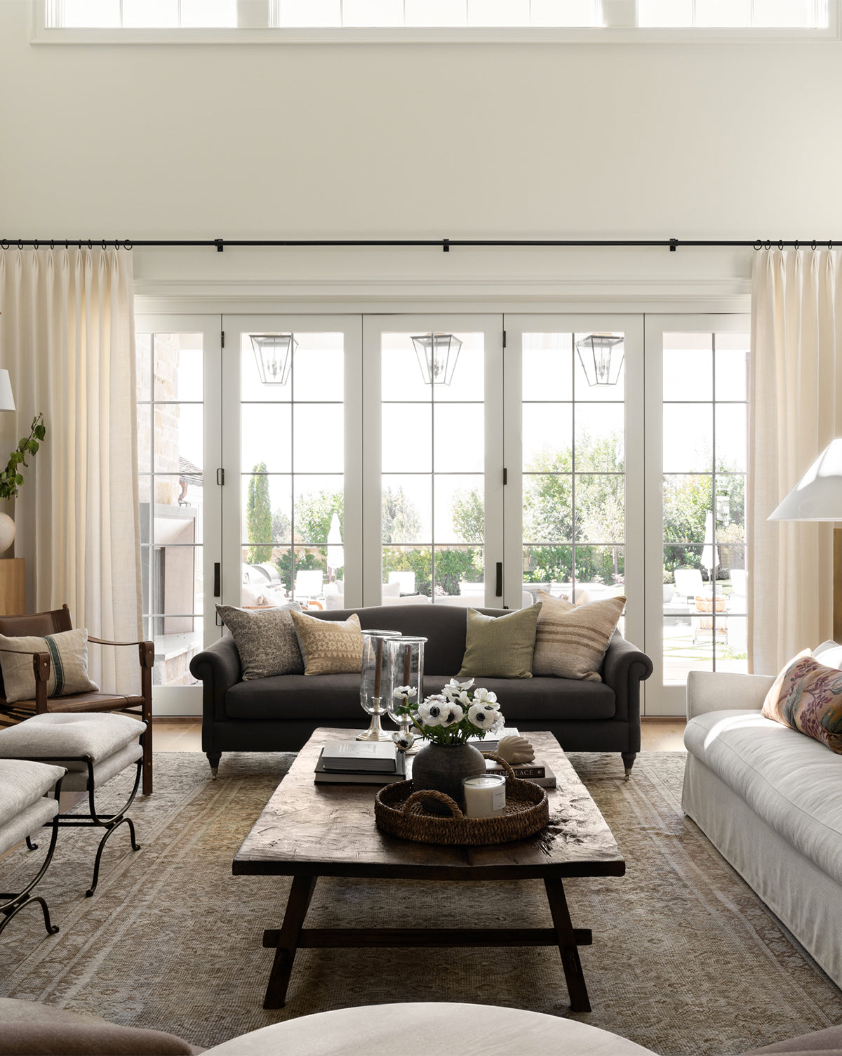 Bright living room with large windows, the customizable Hildegard Sofa 90 in neutral tones, a rustic wooden coffee table with decor, light curtains, and garden patio views. Natural light fills the space, making it warm and inviting by Vendixs