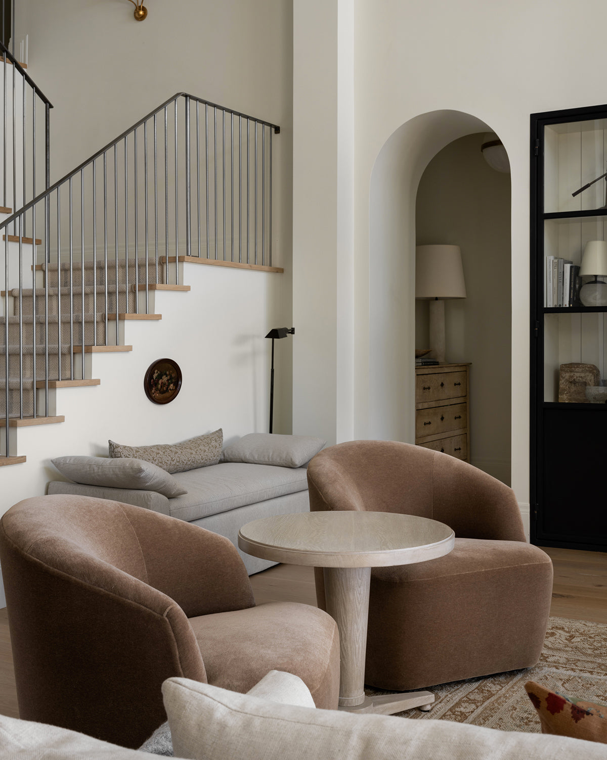 A modern living room features two Makerspalm Byron Chairs in brown paired with a round table, a light gray sofa by the stairs, neutral decor, and a black-framed glass cabinet beside an arched doorway—all thoughtfully styled in collaboration with Vendixs