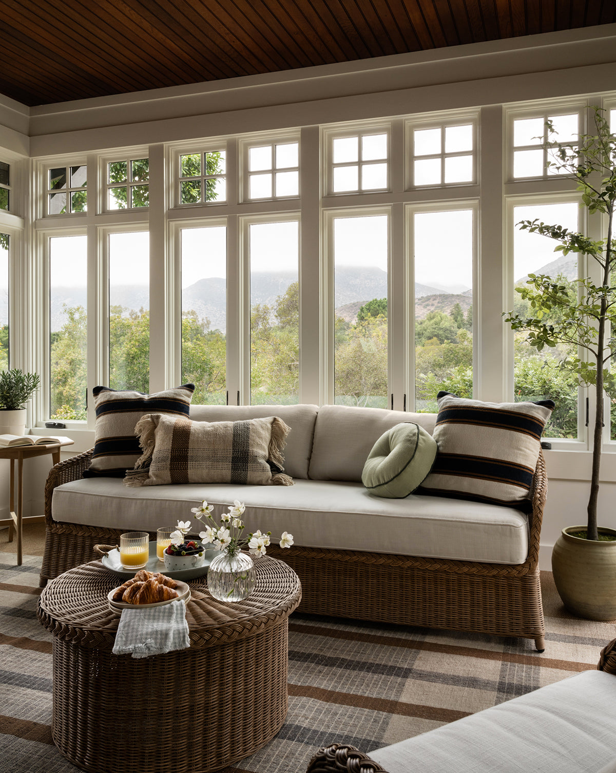 A cozy sunroom with large windows showcases the Haviland Outdoor Sofa 88 with plush pillows and a round wicker coffee table topped with drinks, flowers, and pastries. Greenery and warm natural light enhance this inviting space, by Vendixs
