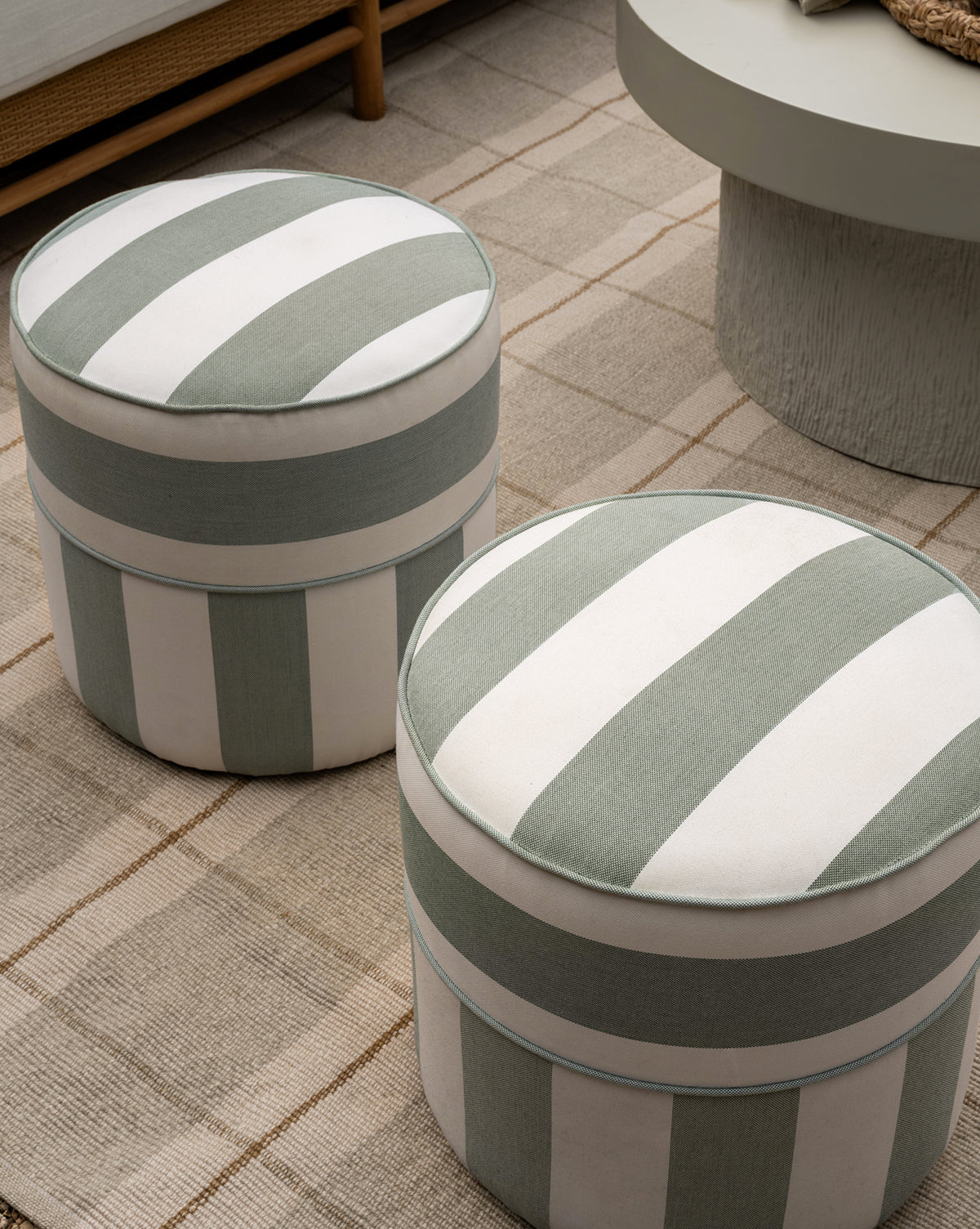 Two Makerspalm Haviland Striped Ottomans with vertical green and white stripes, made for outdoor use, rest on a beige checkered rug in a modern living room next to a round light-gray table. Vendixs