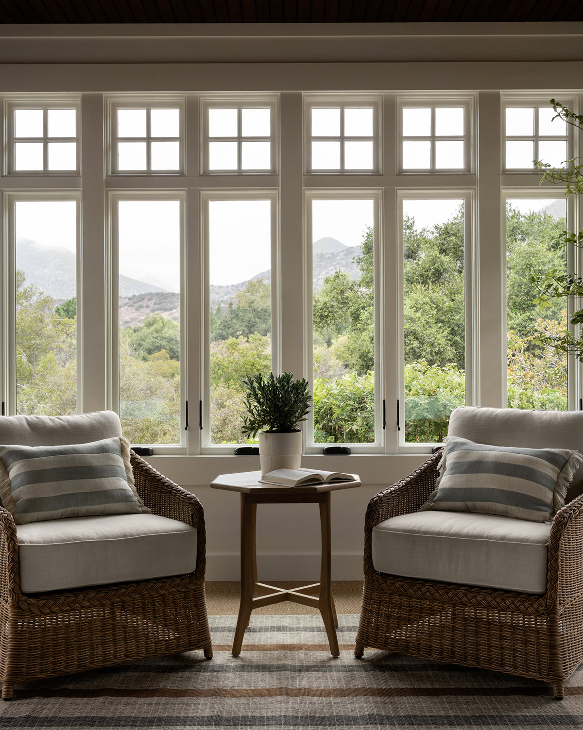 Two Haviland Outdoor Lounge Chairs with striped cushions face each other by a small round table with a plant and book, set in front of large windows with views of trees and distant mountains, by Vendixs