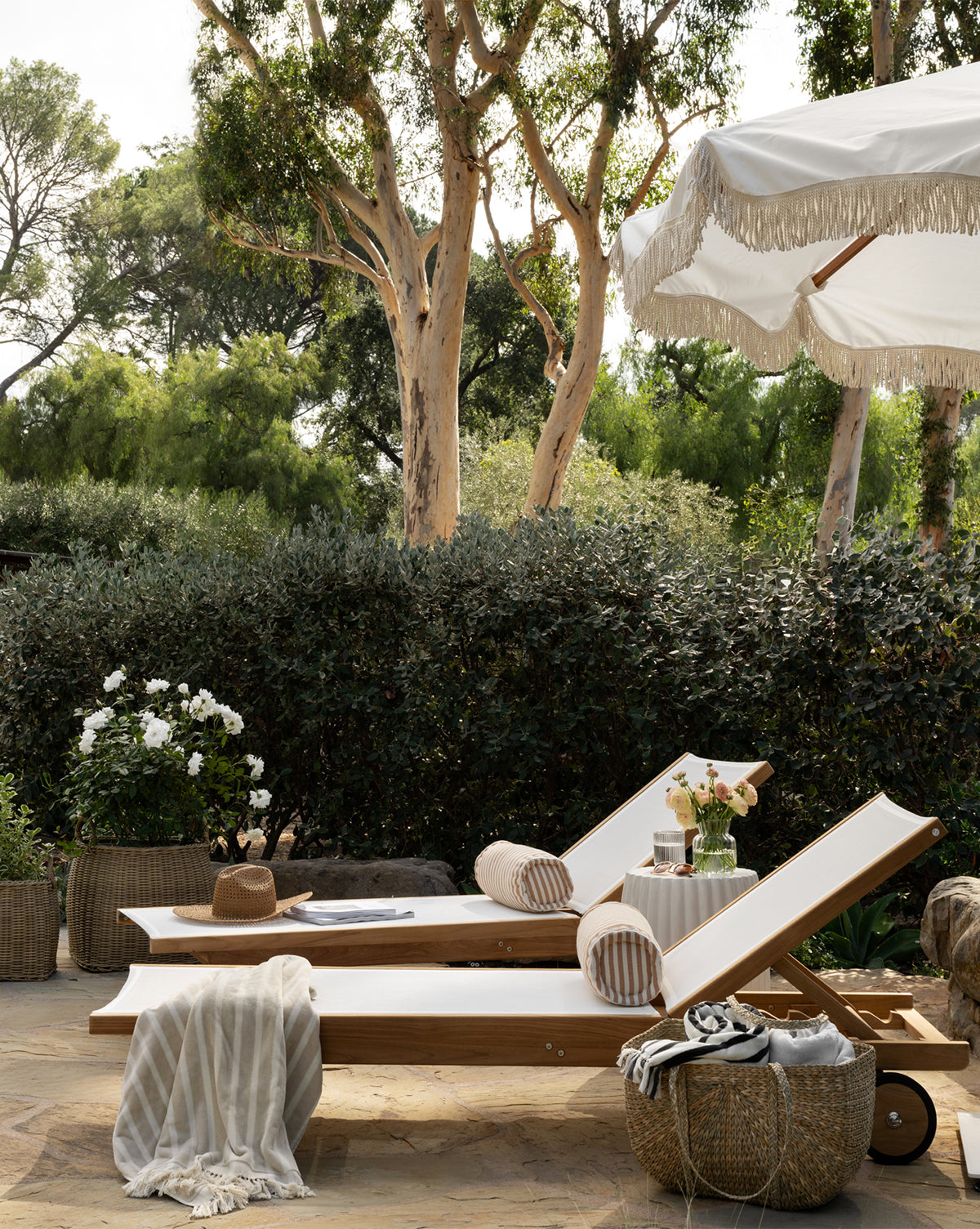 Two Cadie Outdoor Chaise lounge chairs with white, weather-resistant cushions sit side by side outdoors under a fringed umbrella, accented by striped pillows, a hat, blanket, drinks, and a basket amid lush garden greenery by Vendixs