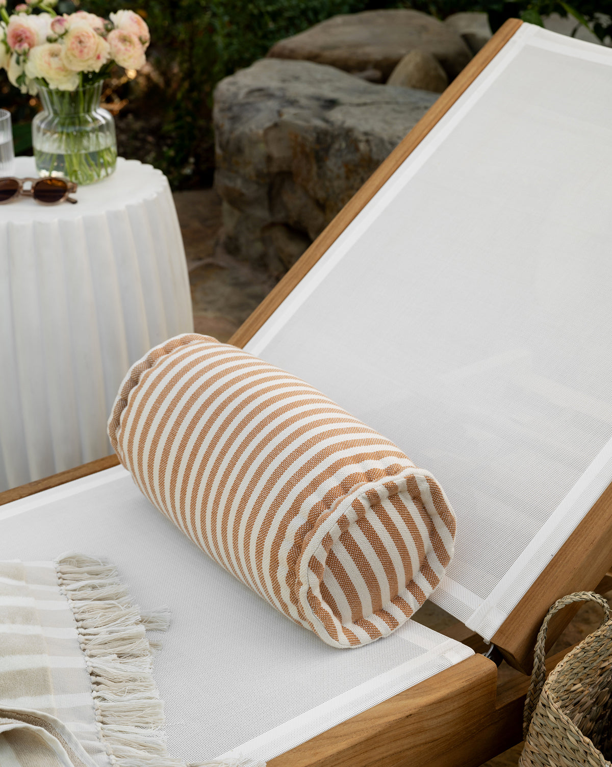 A white lounge chair with a weather-resistant pillow, the Striped Indoor/Outdoor Bolster Pillow in tan and white, and a fringed throw sits by a round table with flowers and sunglasses. Stones and greenery add to the outdoor decor backdrop, by Vendixs