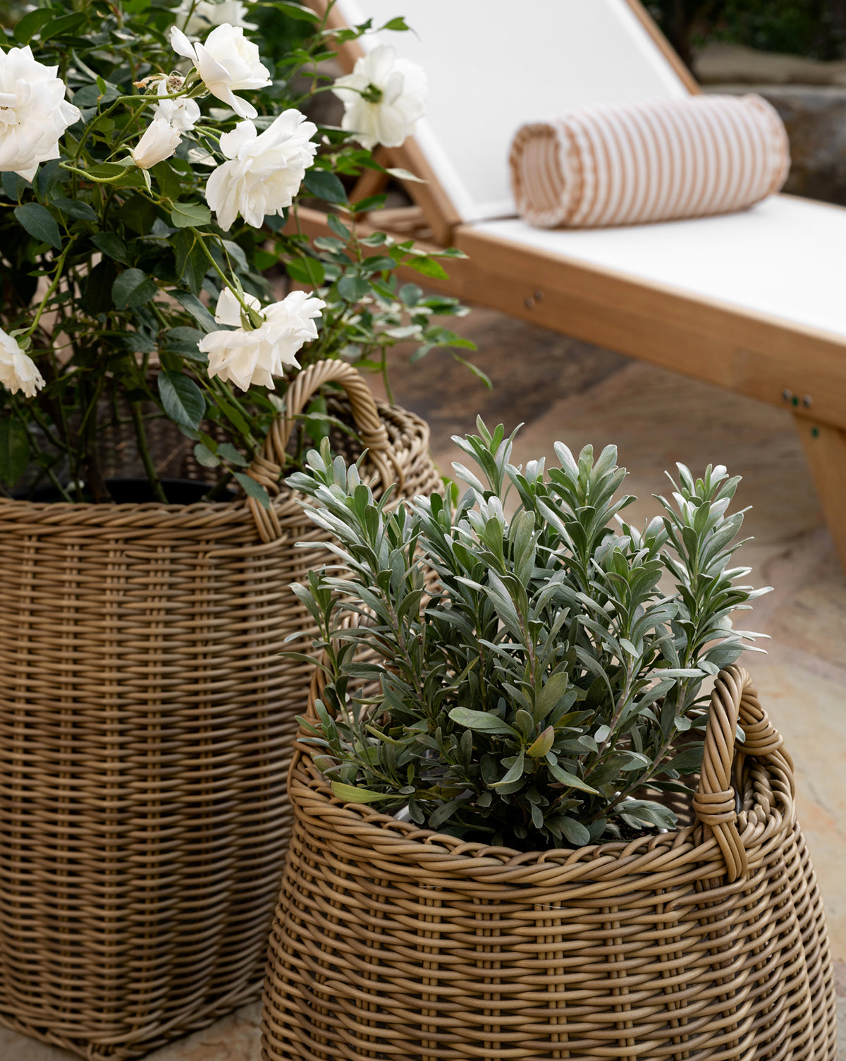 Two Lightwash Handled Planter Baskets with green indoor plants—one blooming white flowers—are placed outdoors beside a wooden lounge chair featuring a white cushion and striped bolster pillow, by Vendixs