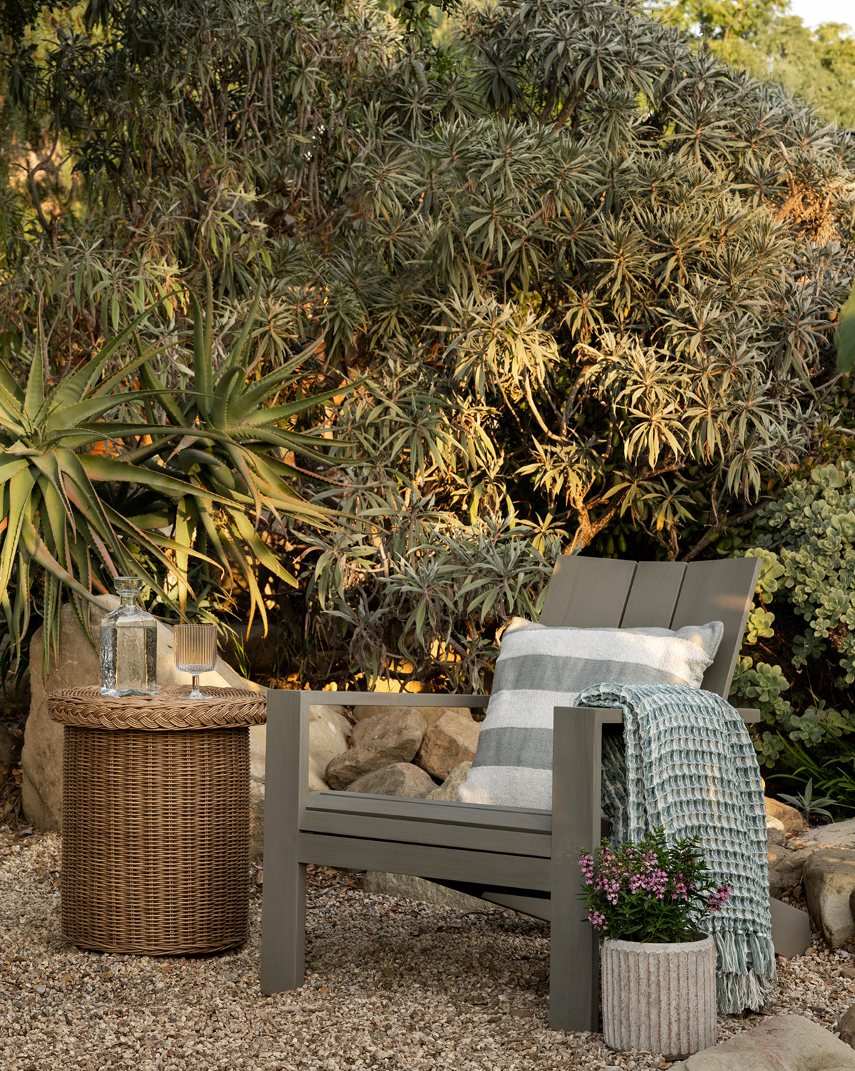 A cozy outdoor lounge featuring a weather-resistant gray wooden chair with striped cushion, blue throw, Haviland Side Table holding a glass water bottle and cup, plus a planter with pink flowers amid lush greenery and gravel, by Vendixs